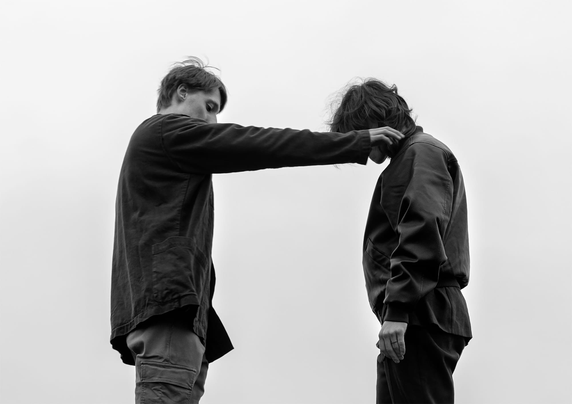 Two people stand facing each other against a gray sky. One extends an arm toward the other's shoulder, creating a tense atmosphere.