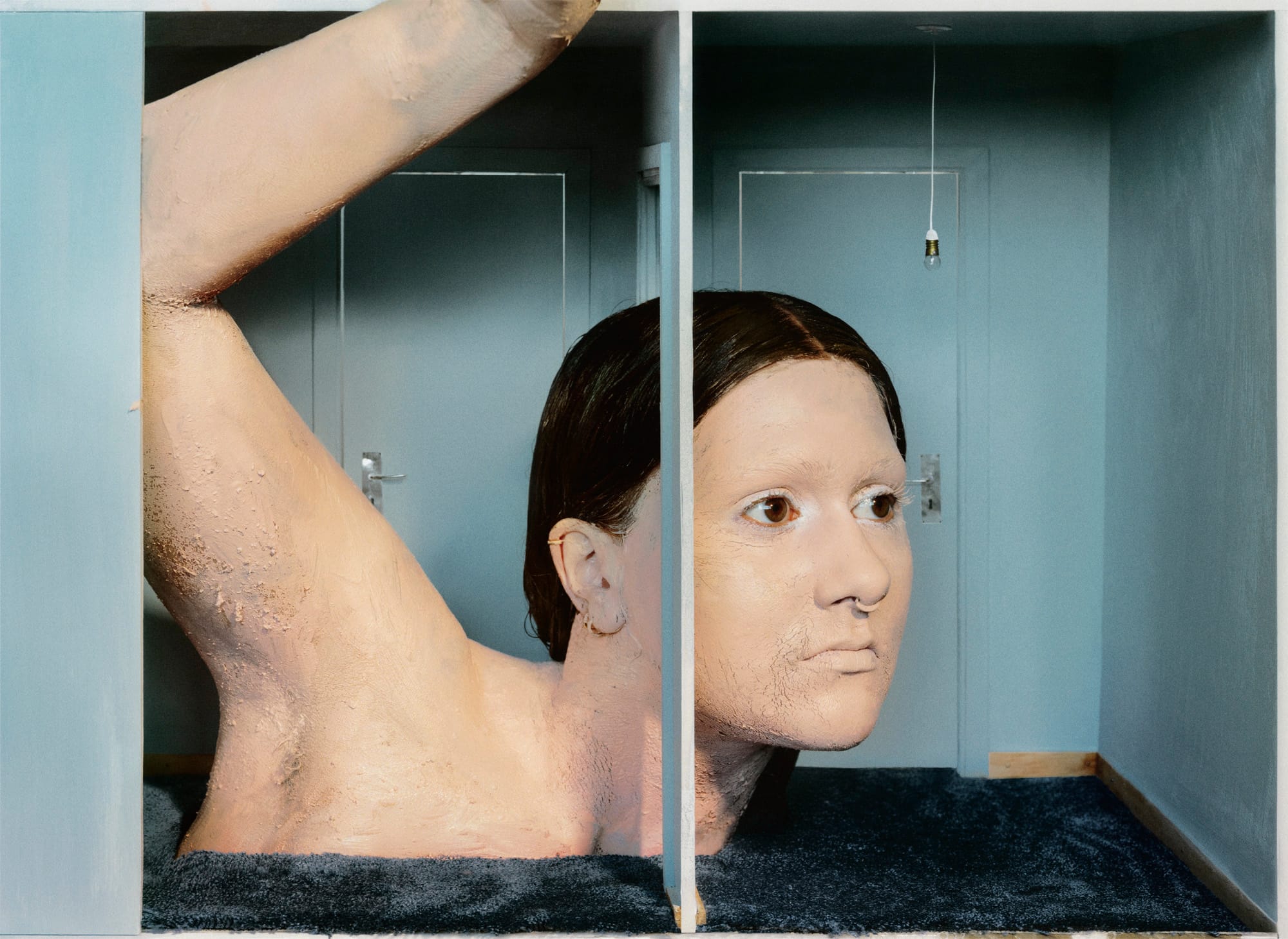A large, textured human face and arm emerge from a blue room, with a single light bulb hanging from the ceiling.