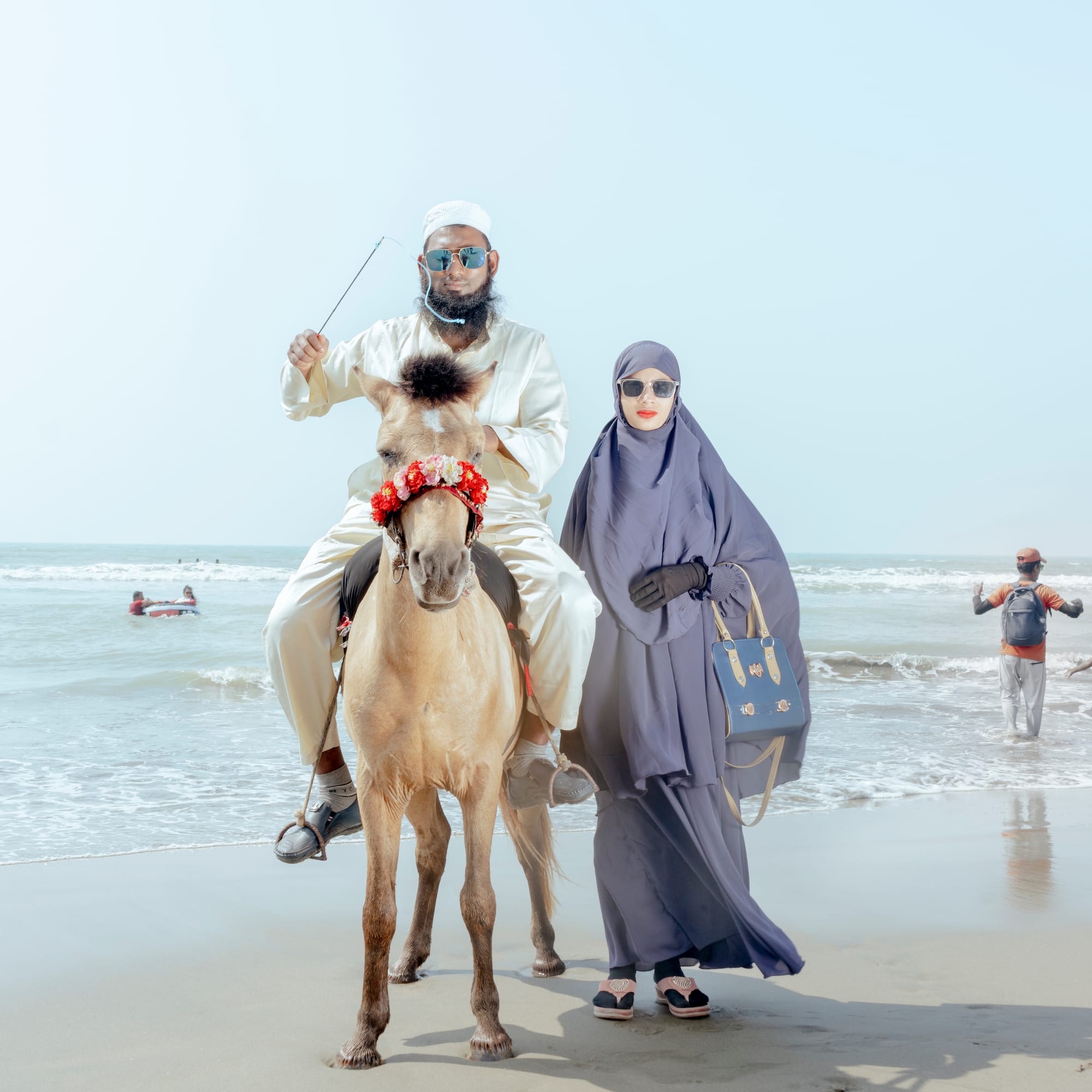 Man in white riding a horse on the beach, holding reins. Woman in blue abaya stands beside him. Ocean waves and people in the background.
