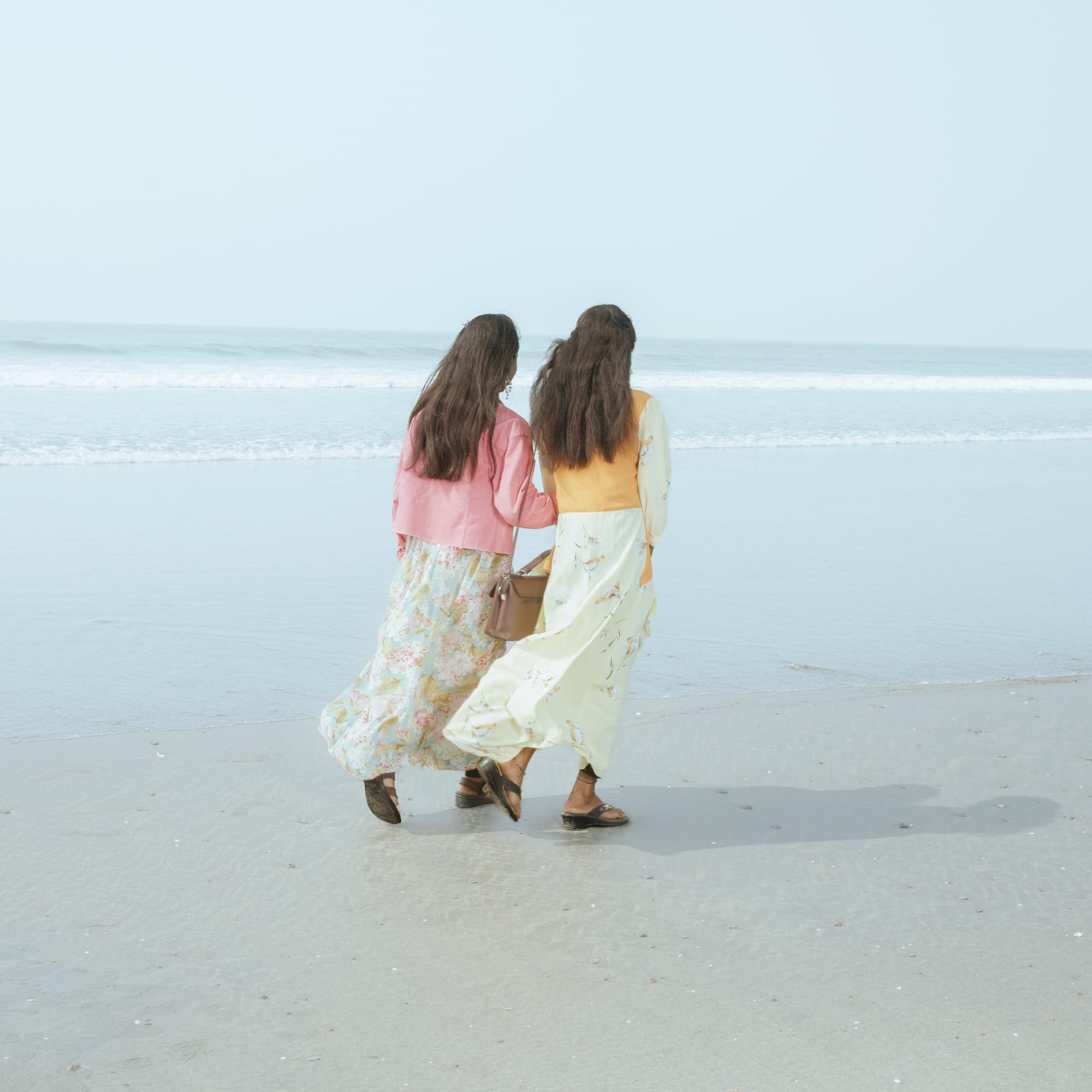 Two women in floral dresses and jackets walk along a sandy beach, facing the ocean.