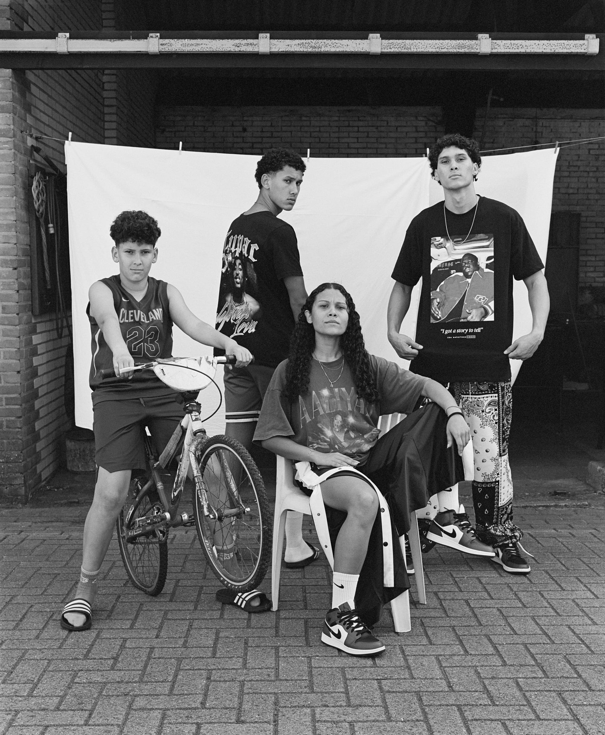 Four people pose confidently in front of a white backdrop. One is on a bike, another is seated, and two stand behind, showcasing bold urban fashion.