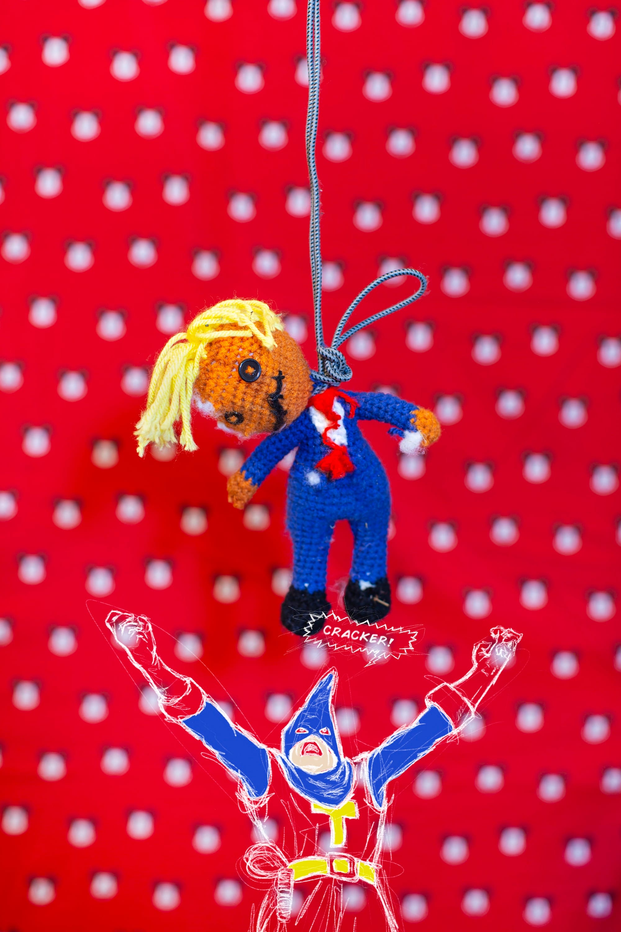 Image of a knitted puppet hanging by its neck on a rope in front of a red dotted background. © André Ramos-Woodard