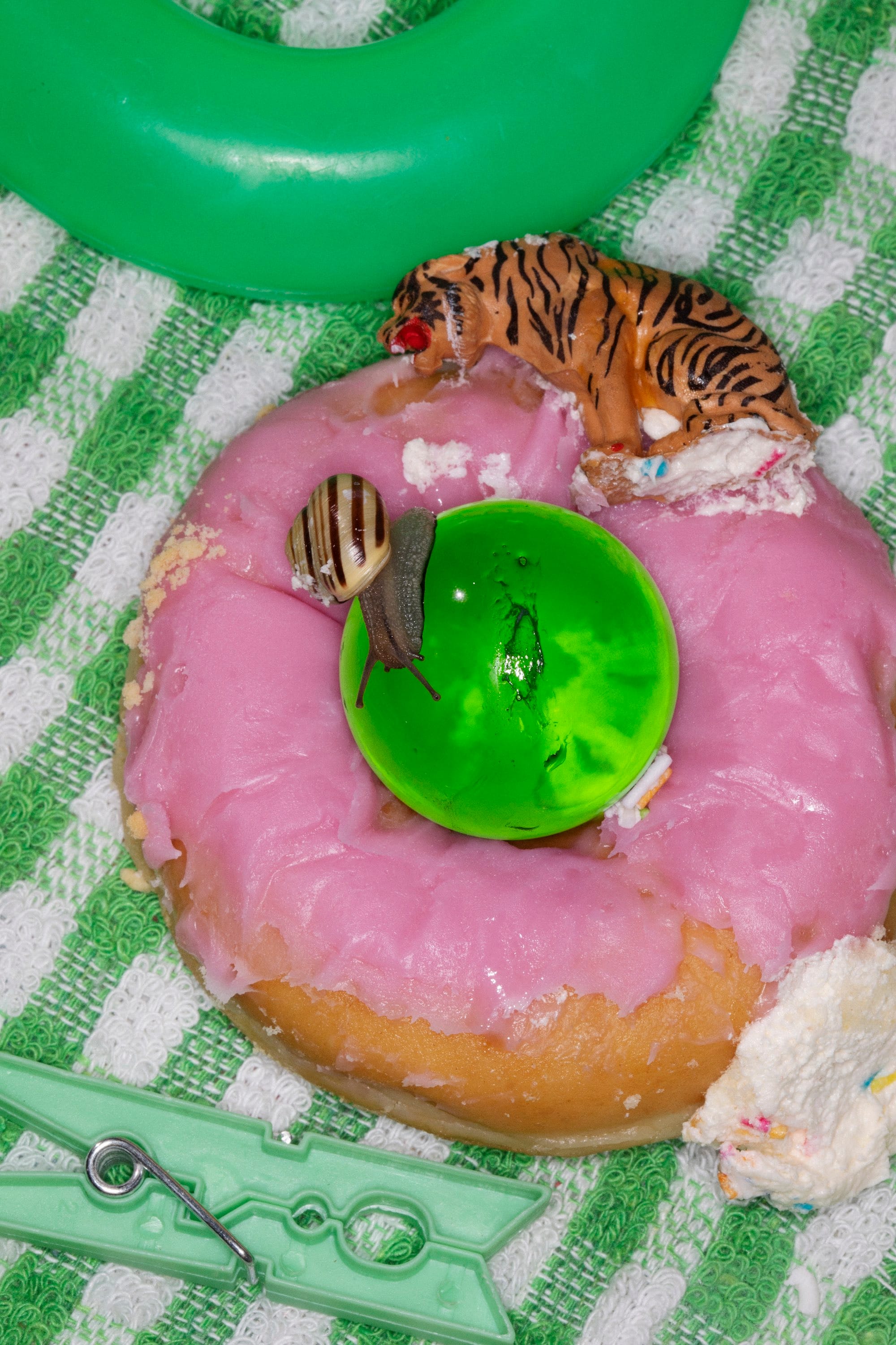 A photograph of a pink-glaced donut, on a green kitchen cloth, surrounded by a green cloth peg, a small toy tiger and a snail.