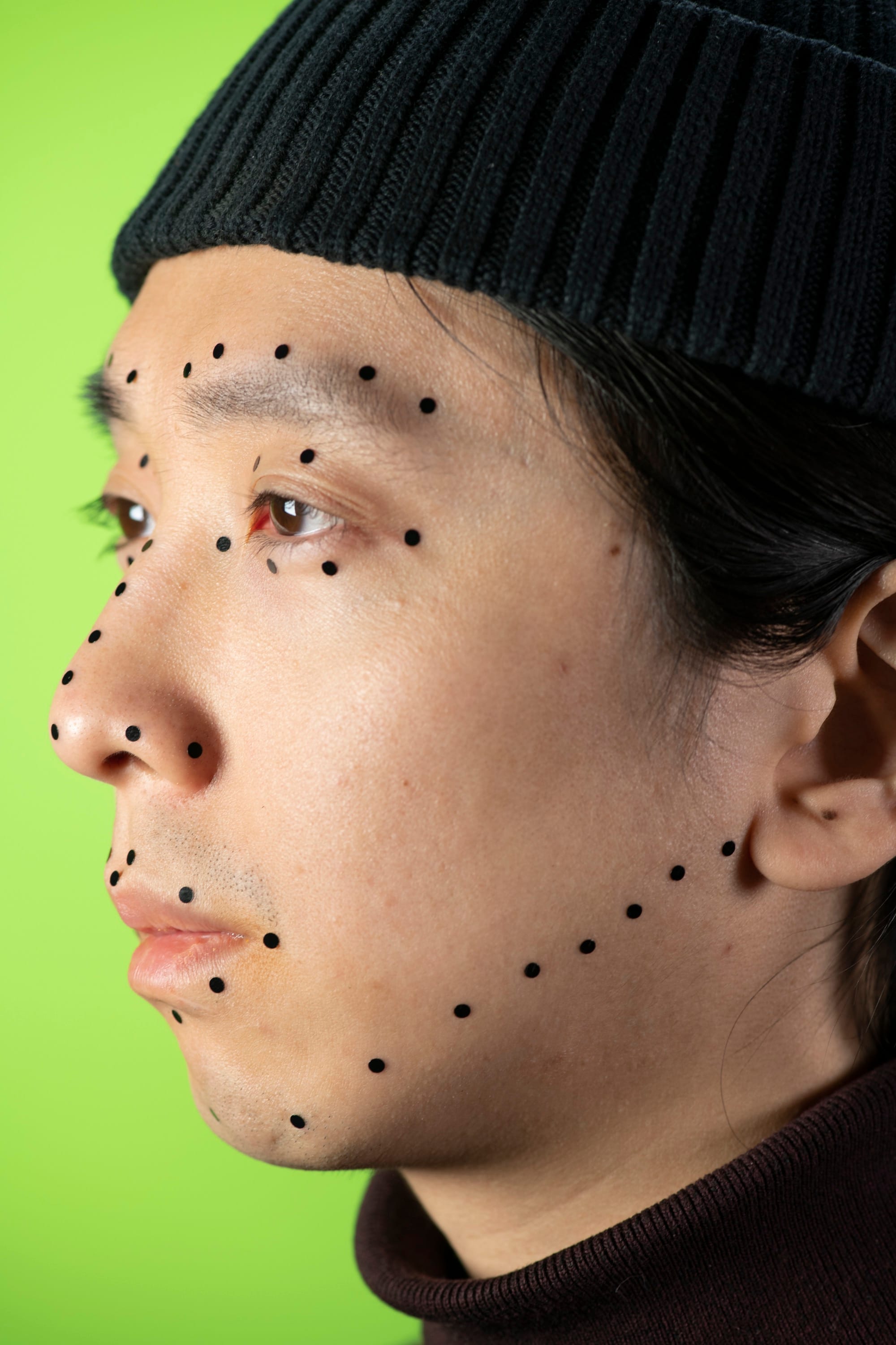 Portrait of the artist in profile in front of a green background, wearing a black hat. His face is dotted with black dots © Sheung Yiu