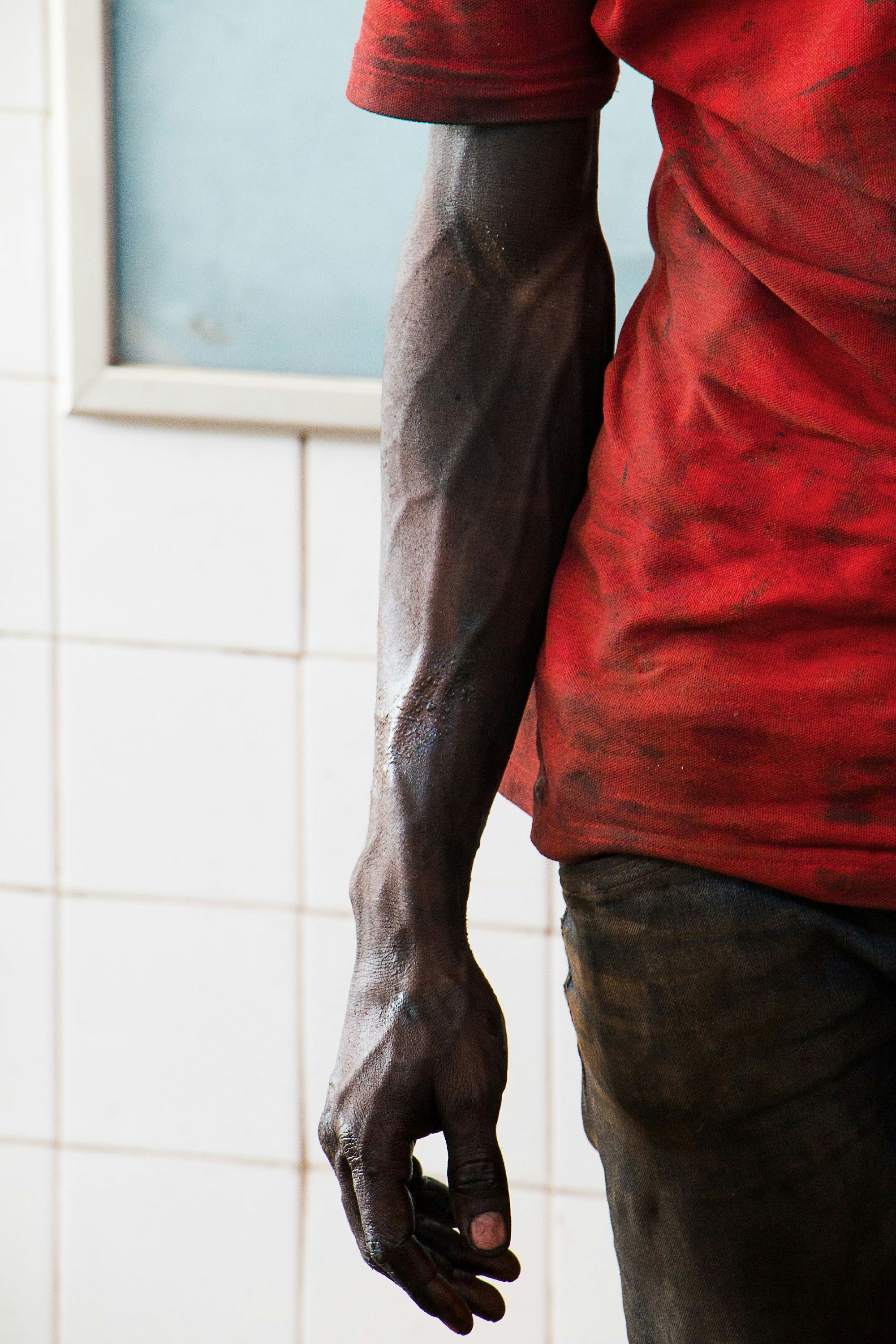 Cropped image of a black man's arm with visible veins and a red t-shirt in front of a tiled wall.