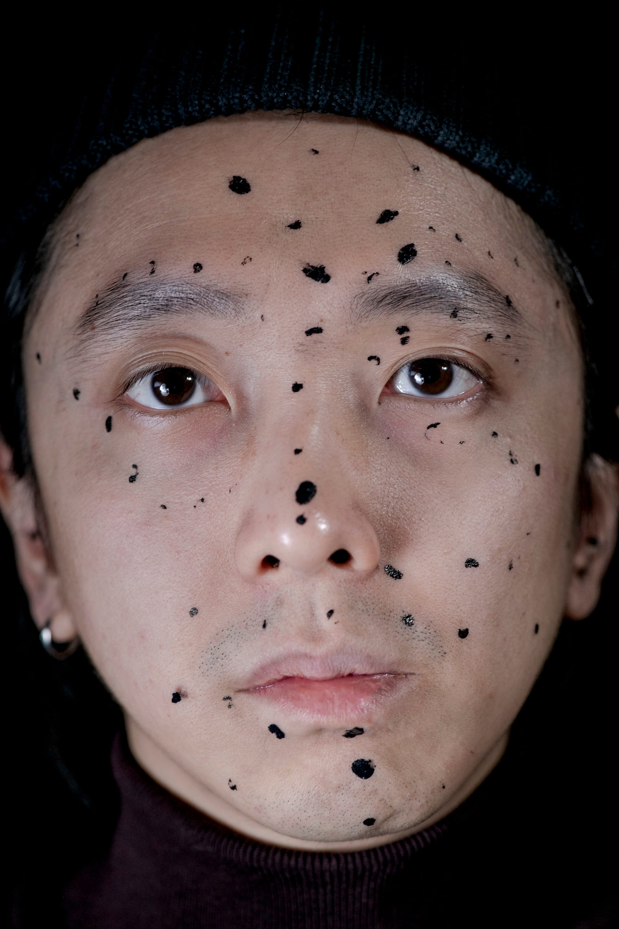 Frontal portrait of the artist's face, his face marked by black paint dots of different shapes and sizes© Sheung Yiu