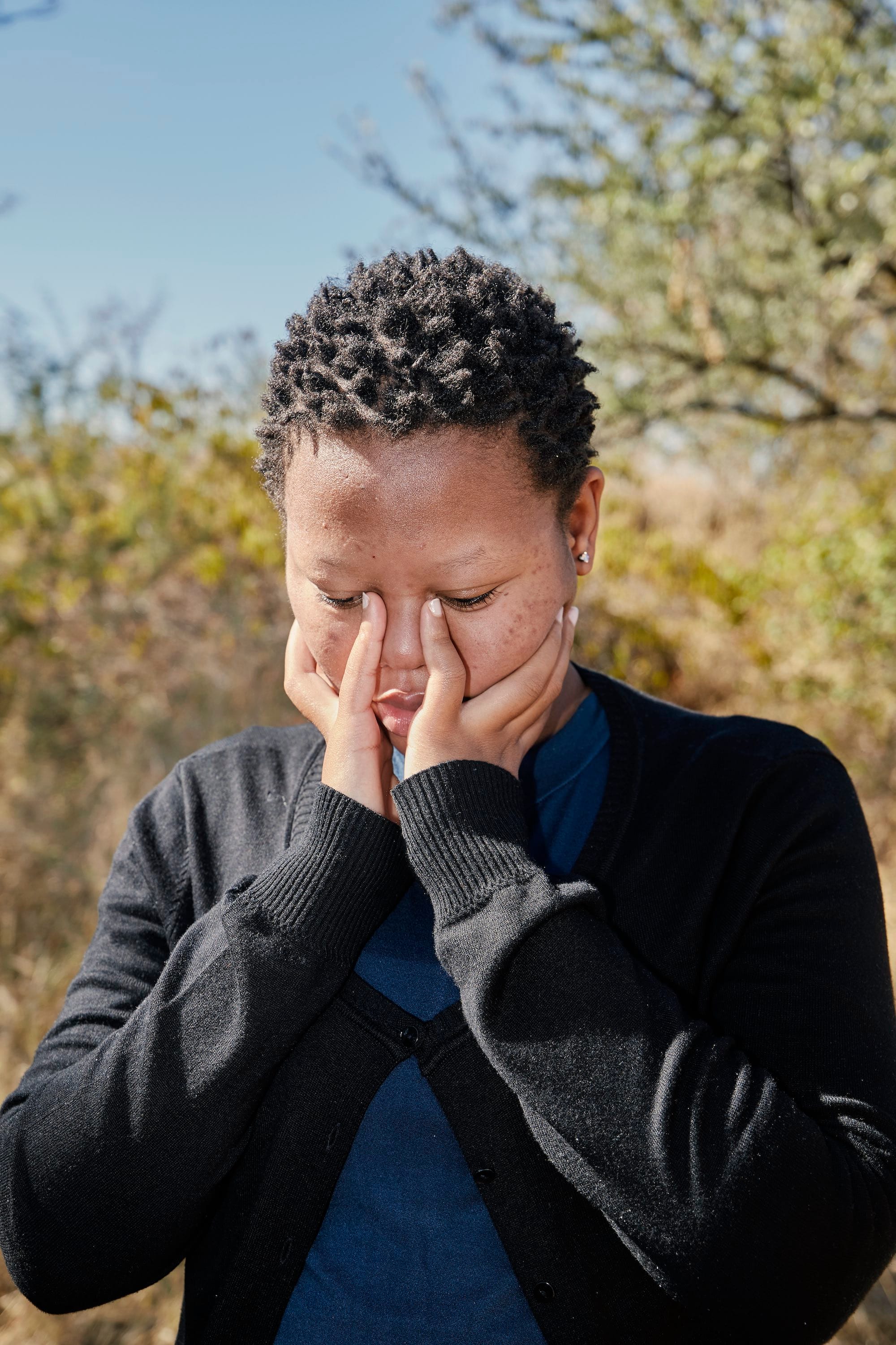 Portrait of a young Black woman in a black cardigan, holding her face in her hands© Thero Makepe