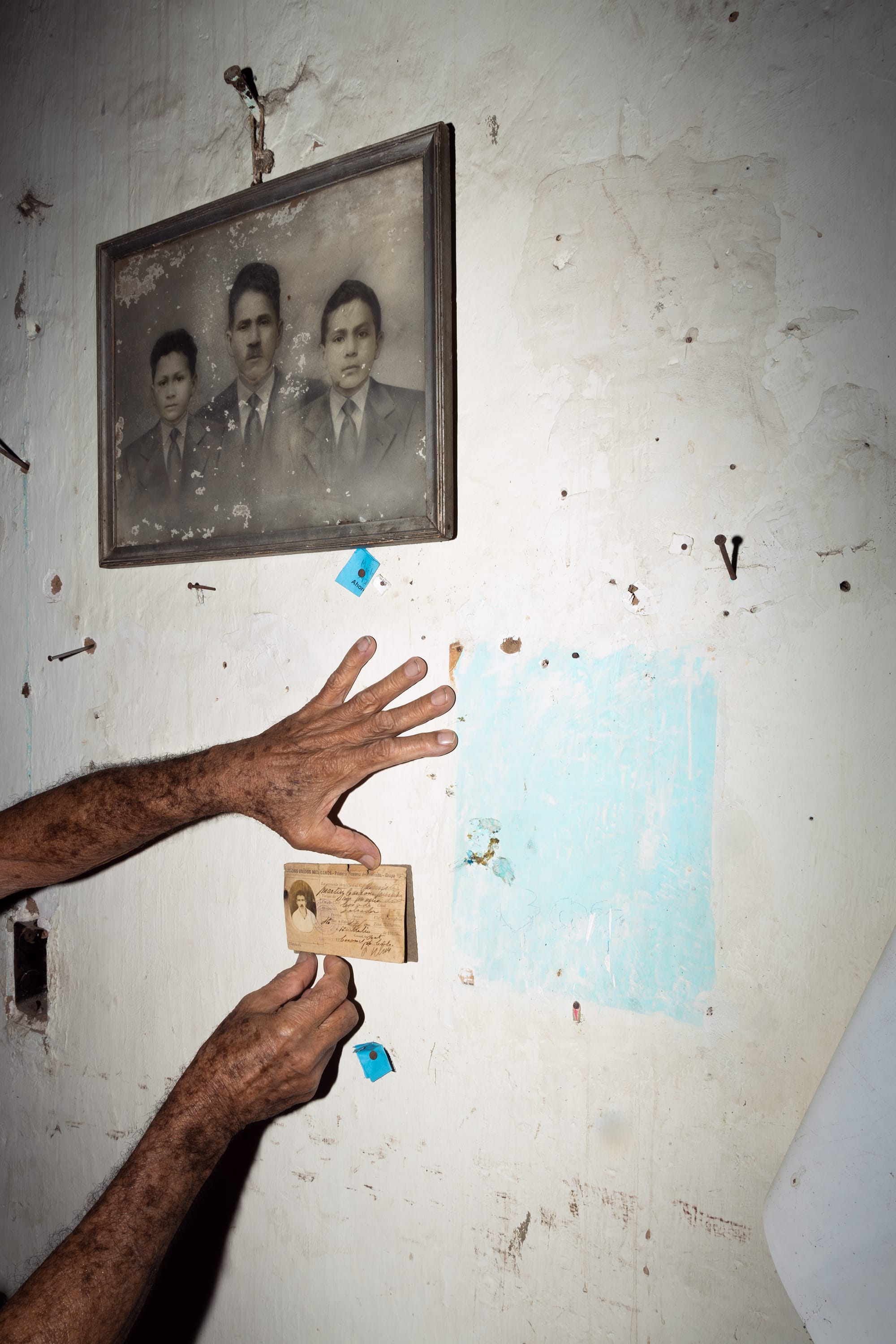 An aged hand holds a small photograph against a worn wall; a framed vintage portrait of three men hangs above.