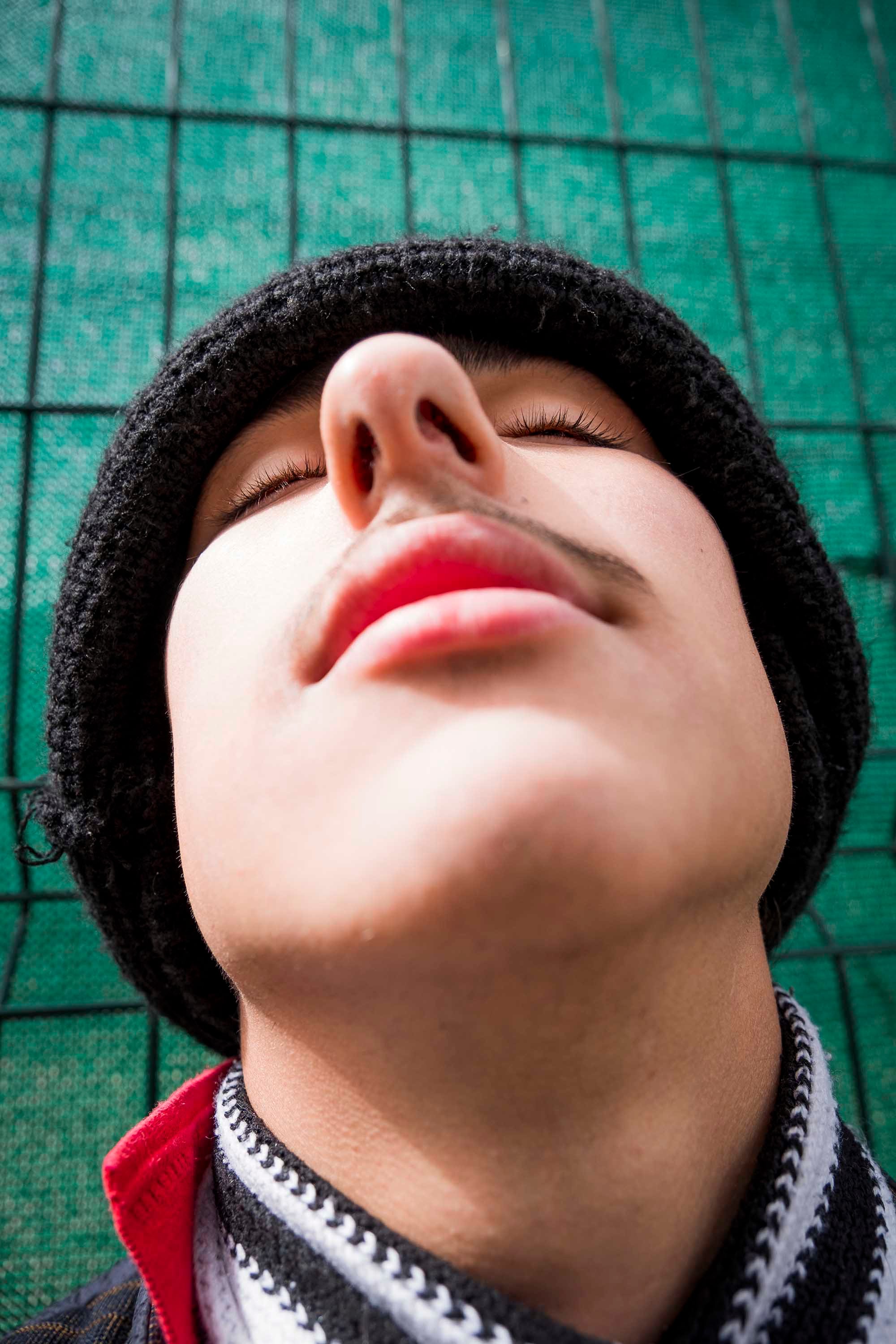 Portrait of a boy wearing a black hat in front of a green gate, with his eyes closed looking up. © Oğulcan Arslan