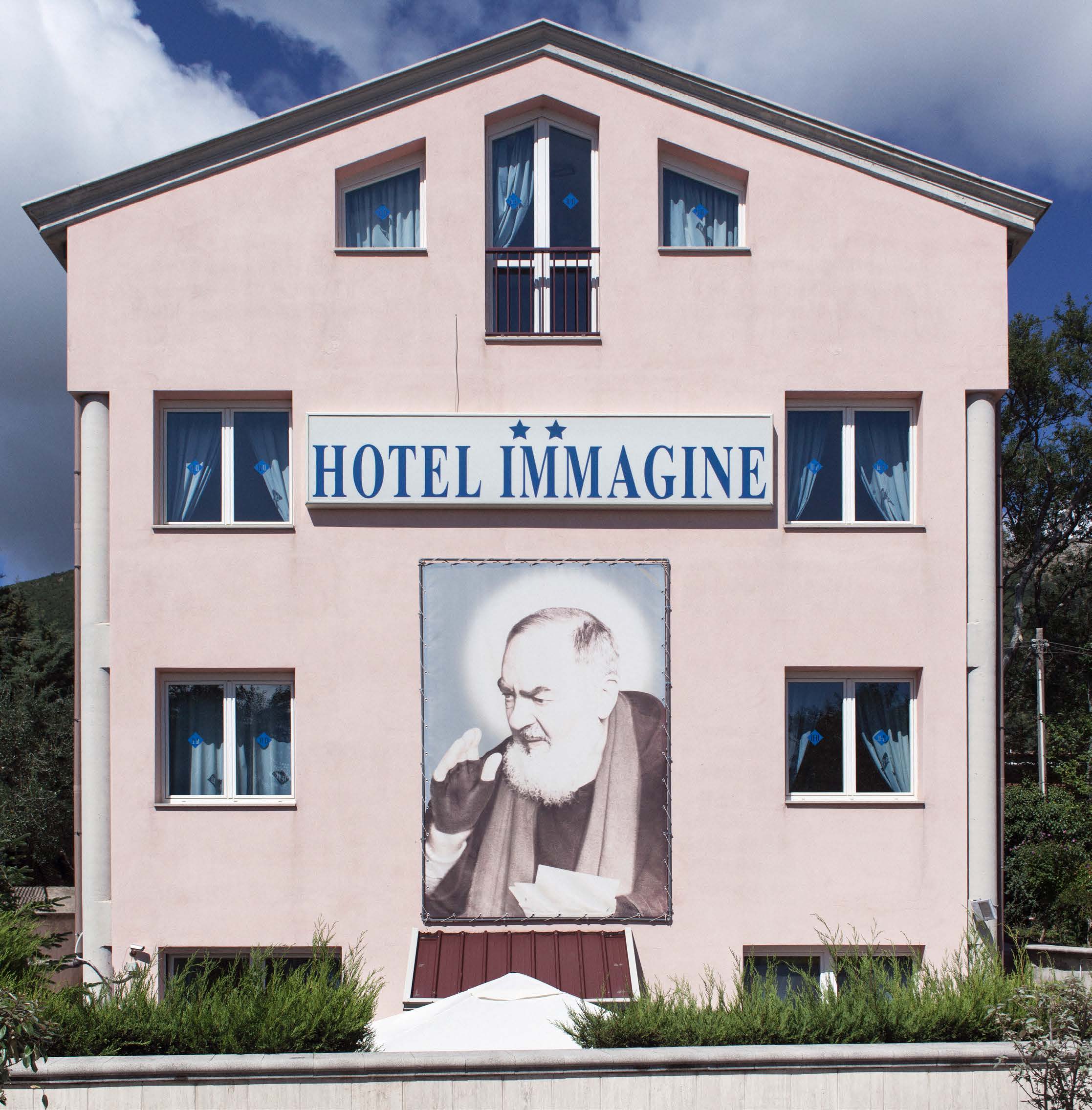 Pink three-story hotel with "Hotel Immagine" sign and large portrait of a bearded man on the facade, surrounded by trees and cloudy sky.
