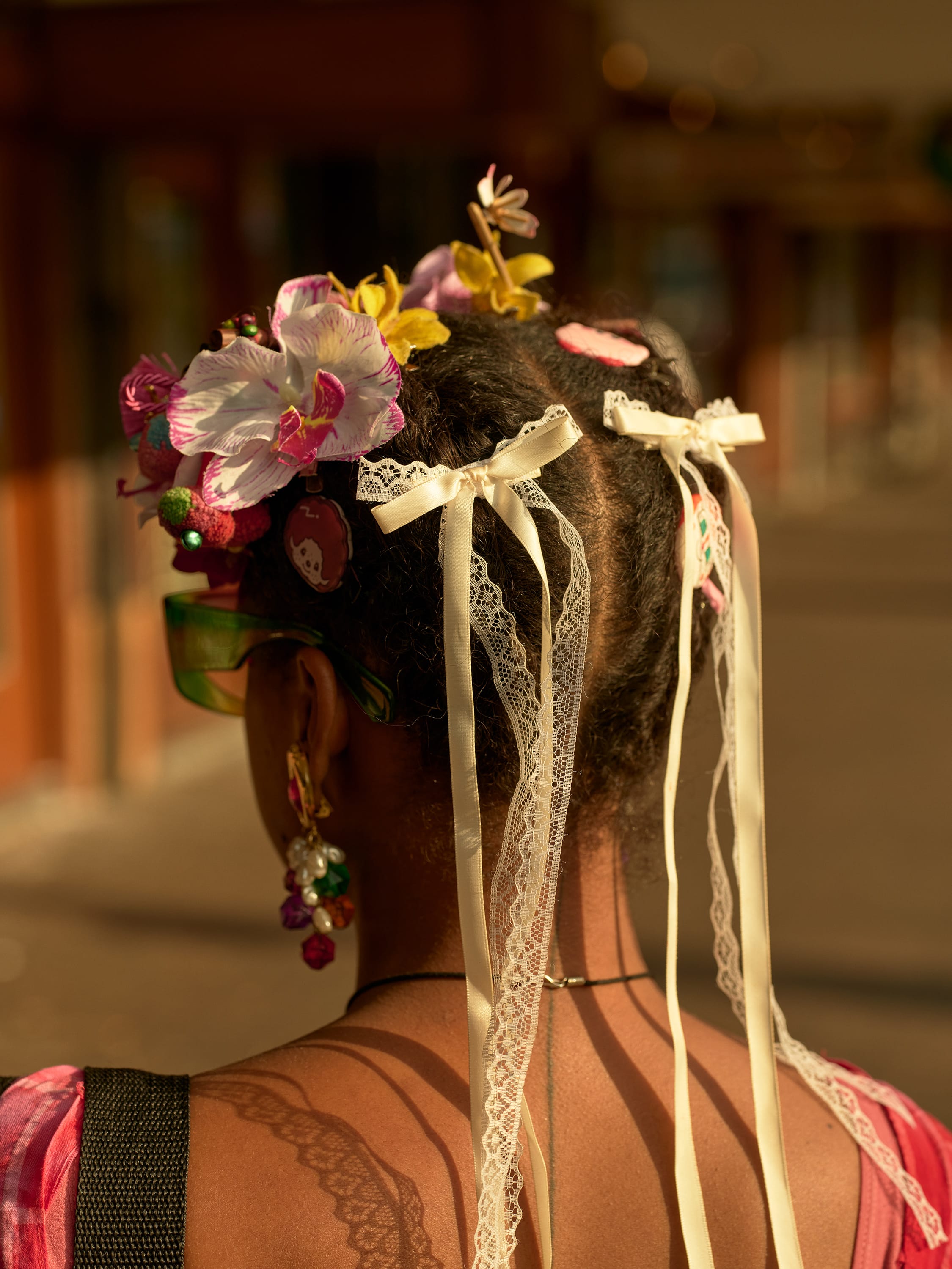 Person with braided hair adorned with colorful flowers, pins, and lace ribbons, wearing large green sunglasses and vibrant earrings.