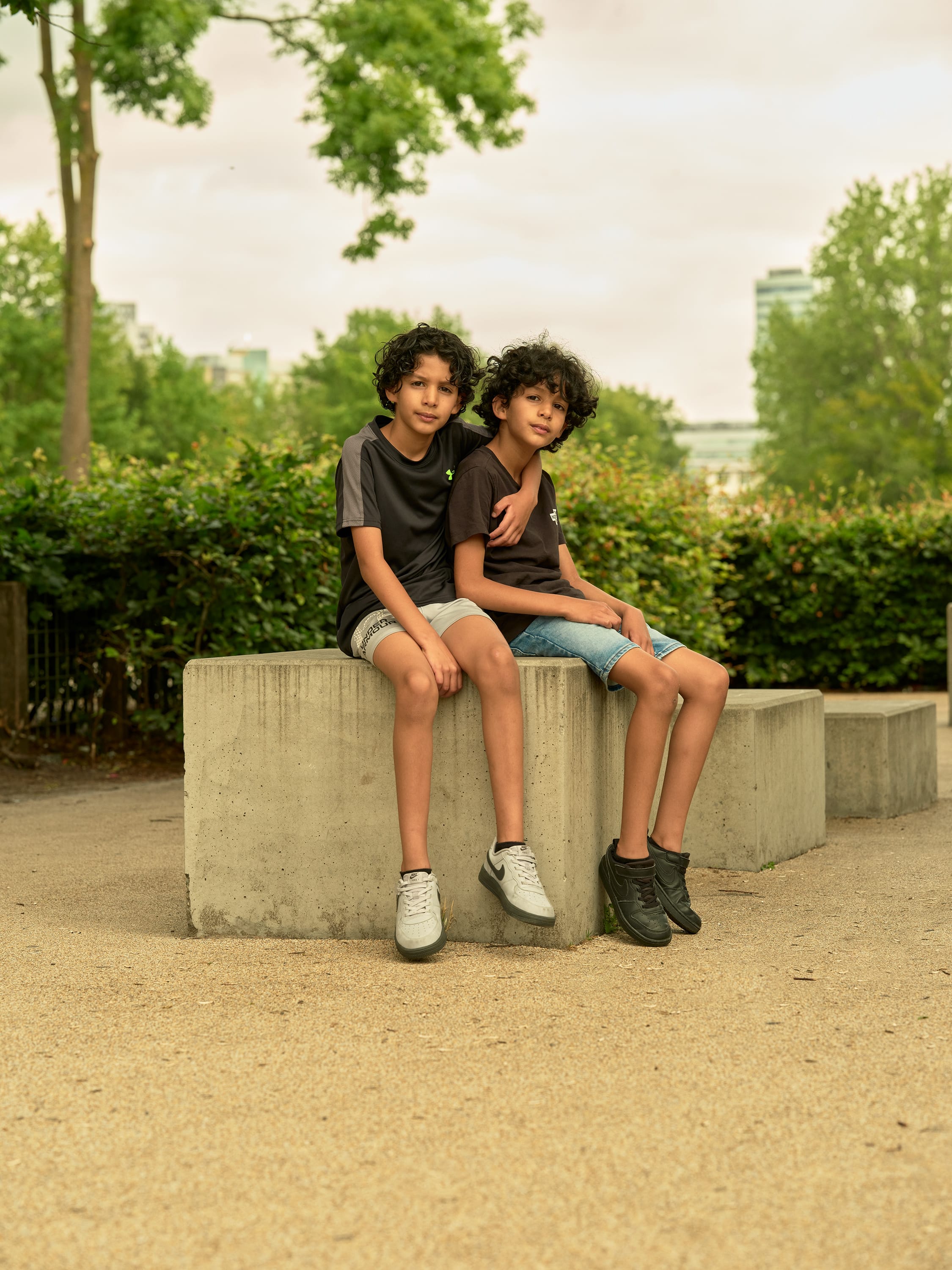 Two boys with curly hair sit on a concrete block in a park, one with an arm around the other. Greenery and trees are in the background.
