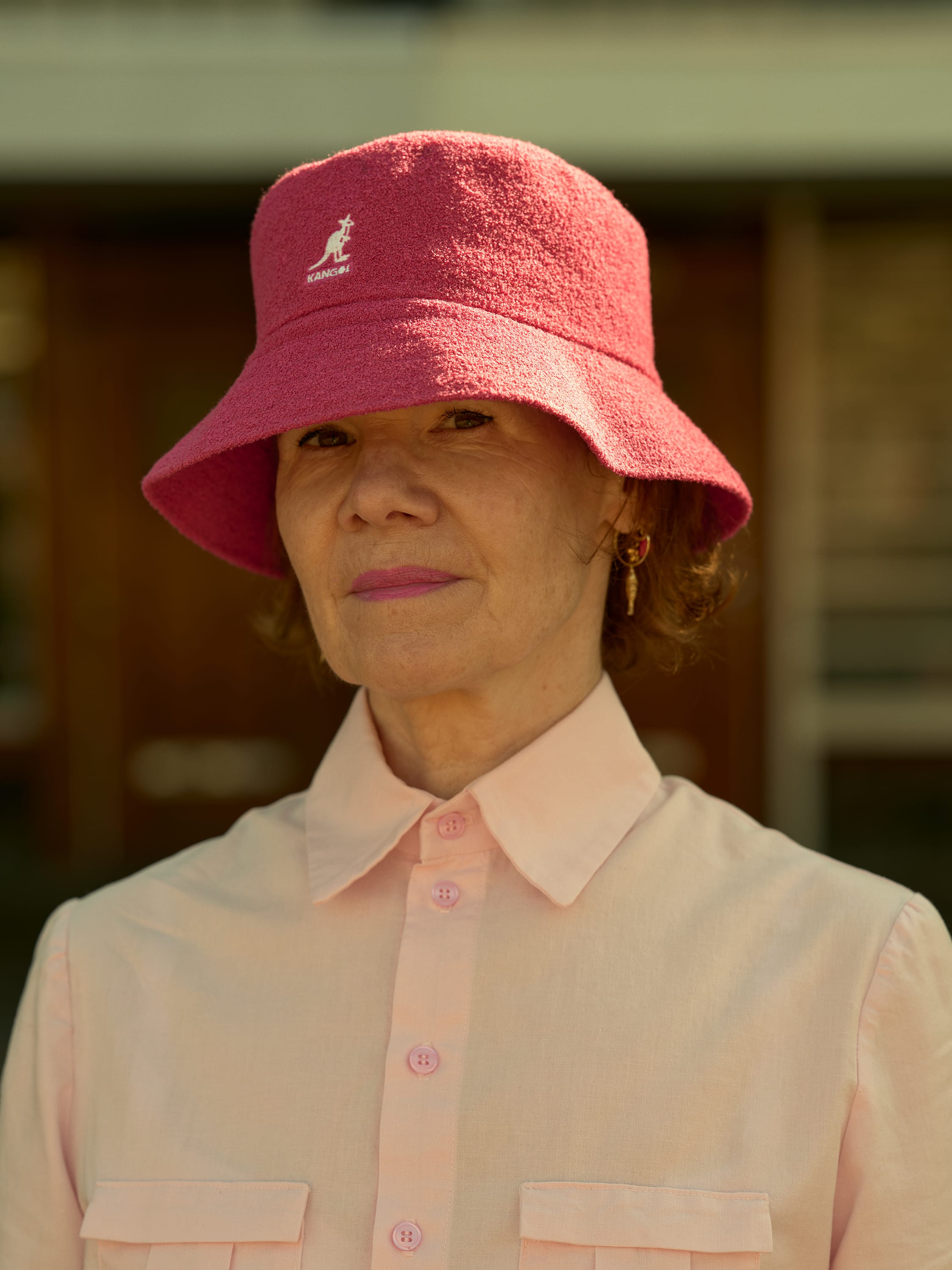 Woman in a pink shirt and matching bucket hat stands outdoors, with a neutral expression and blurred background.
