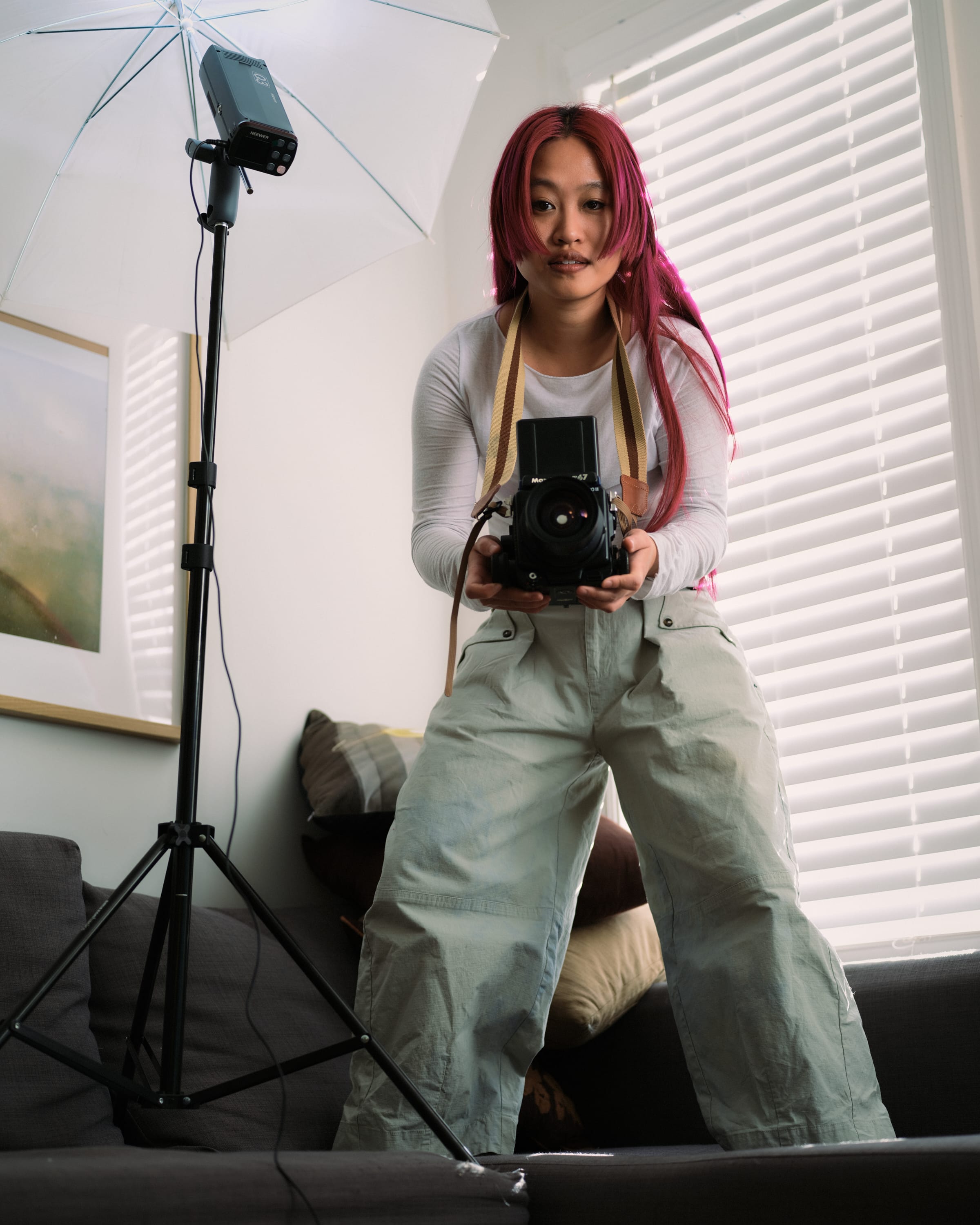 Person with pink hair holds a camera, standing in a room with studio lighting equipment and blinds in the background.