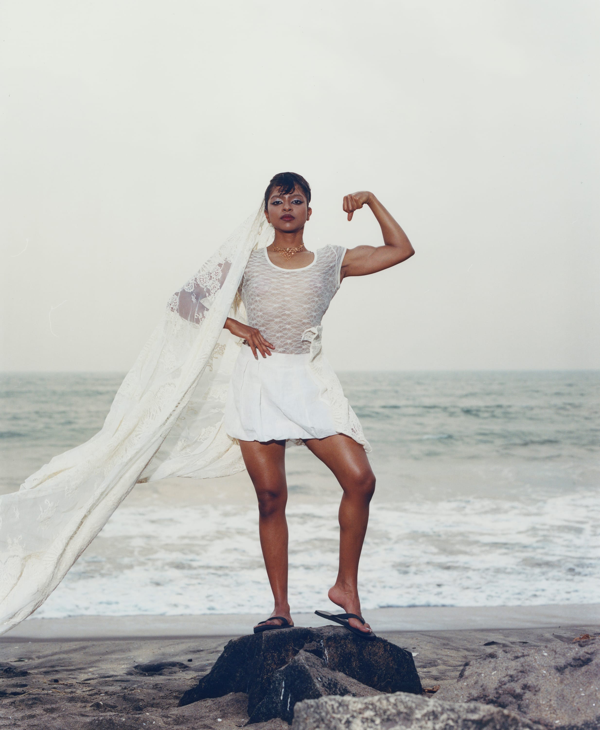 Person in white outfit poses confidently on a rock by the ocean, with a flowing veil and flexed arm, against a cloudy sky.