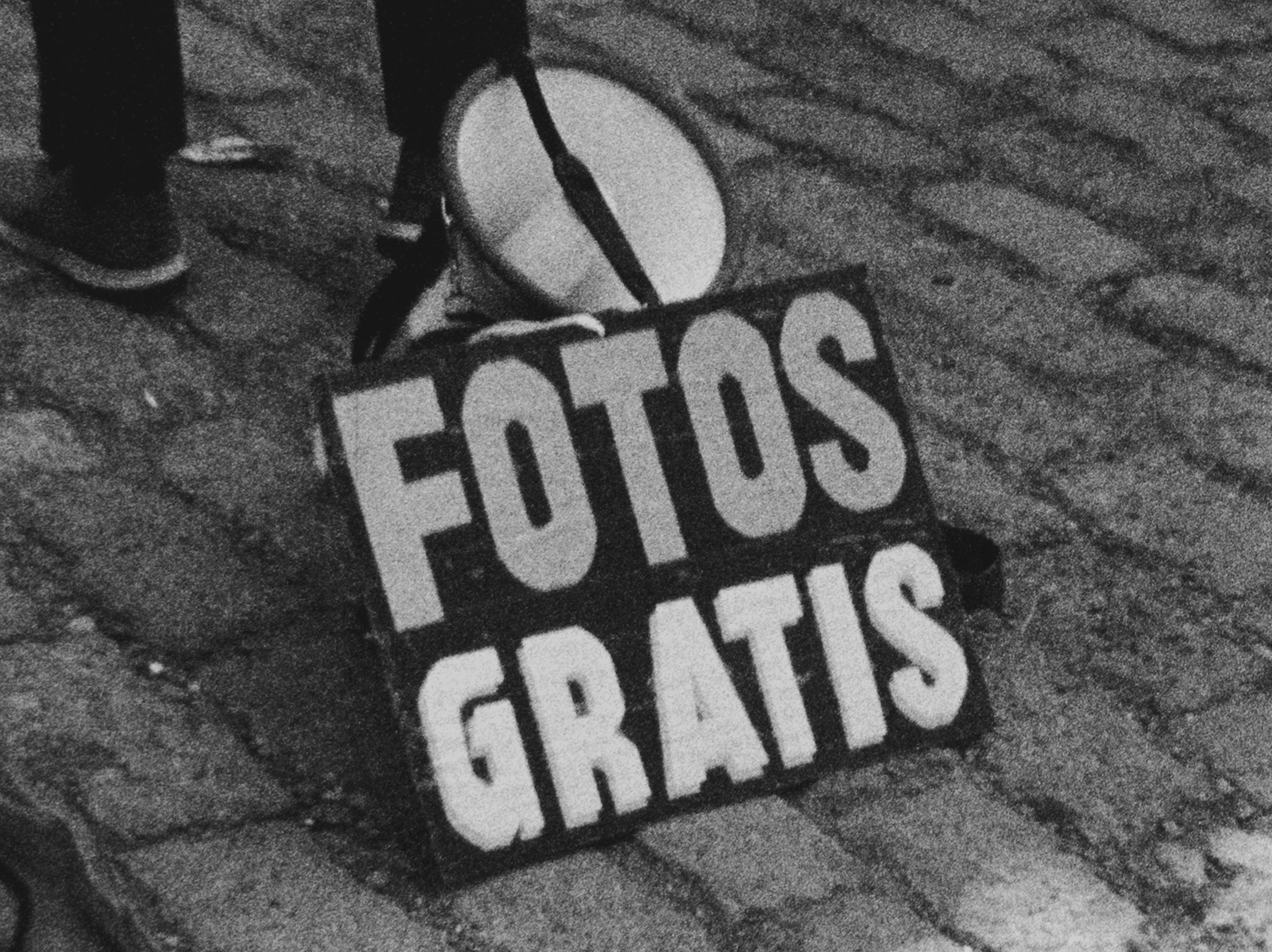 Black and white photo of a sign on cobblestones reading "FOTOS GRATIS" next to a megaphone and a person's leg.