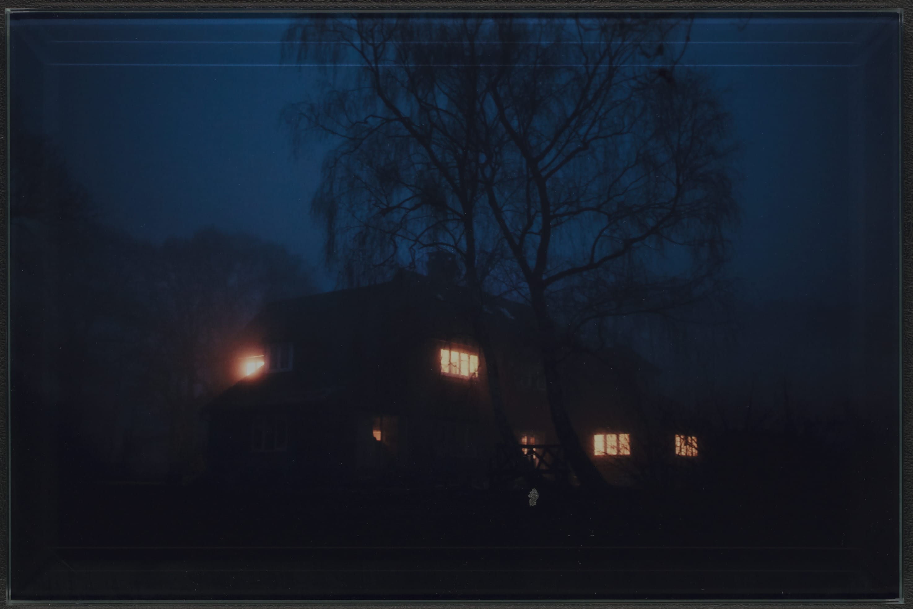 Een huis in het donker met verlichte ramen, twee grote bomen staan voor het huis.