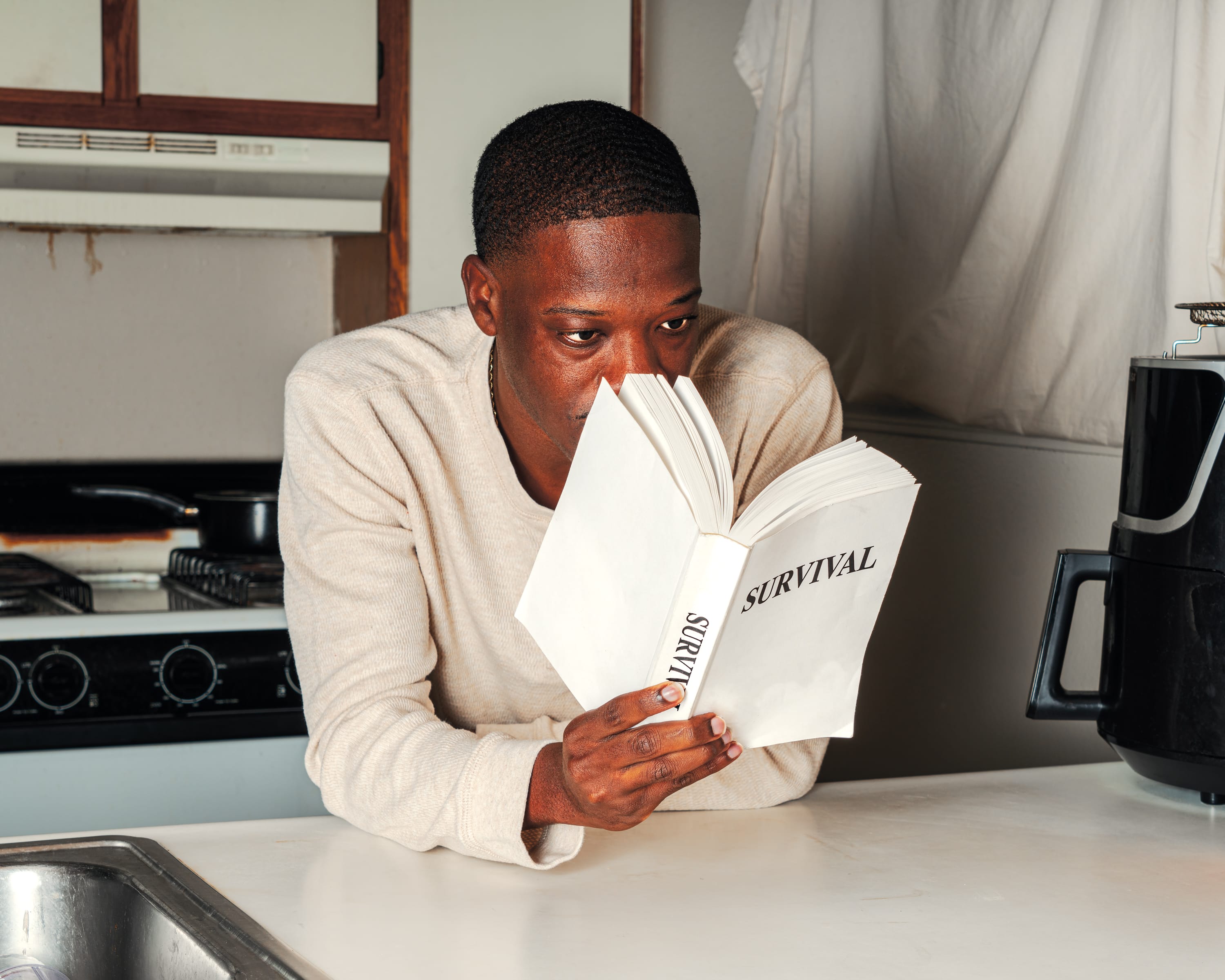 Man in a kitchen intently reading a book titled "Survival" while leaning on a counter near a stove and coffee machine.
