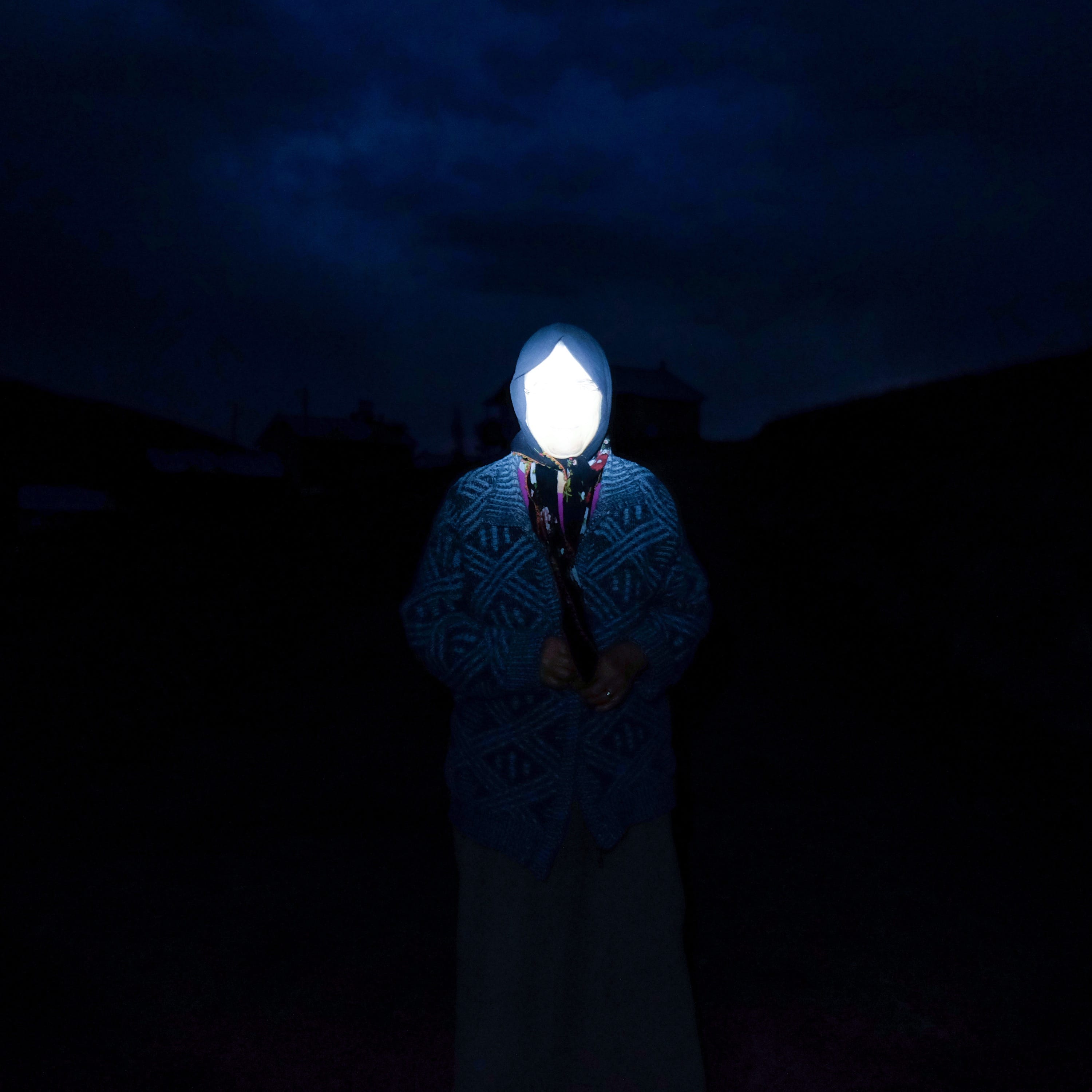 Portrait of a woman in the Kushmer Highlands at night, wearing a head scarf. Her face is invisible due to the flash of the camera ©Cansu Yıldıran