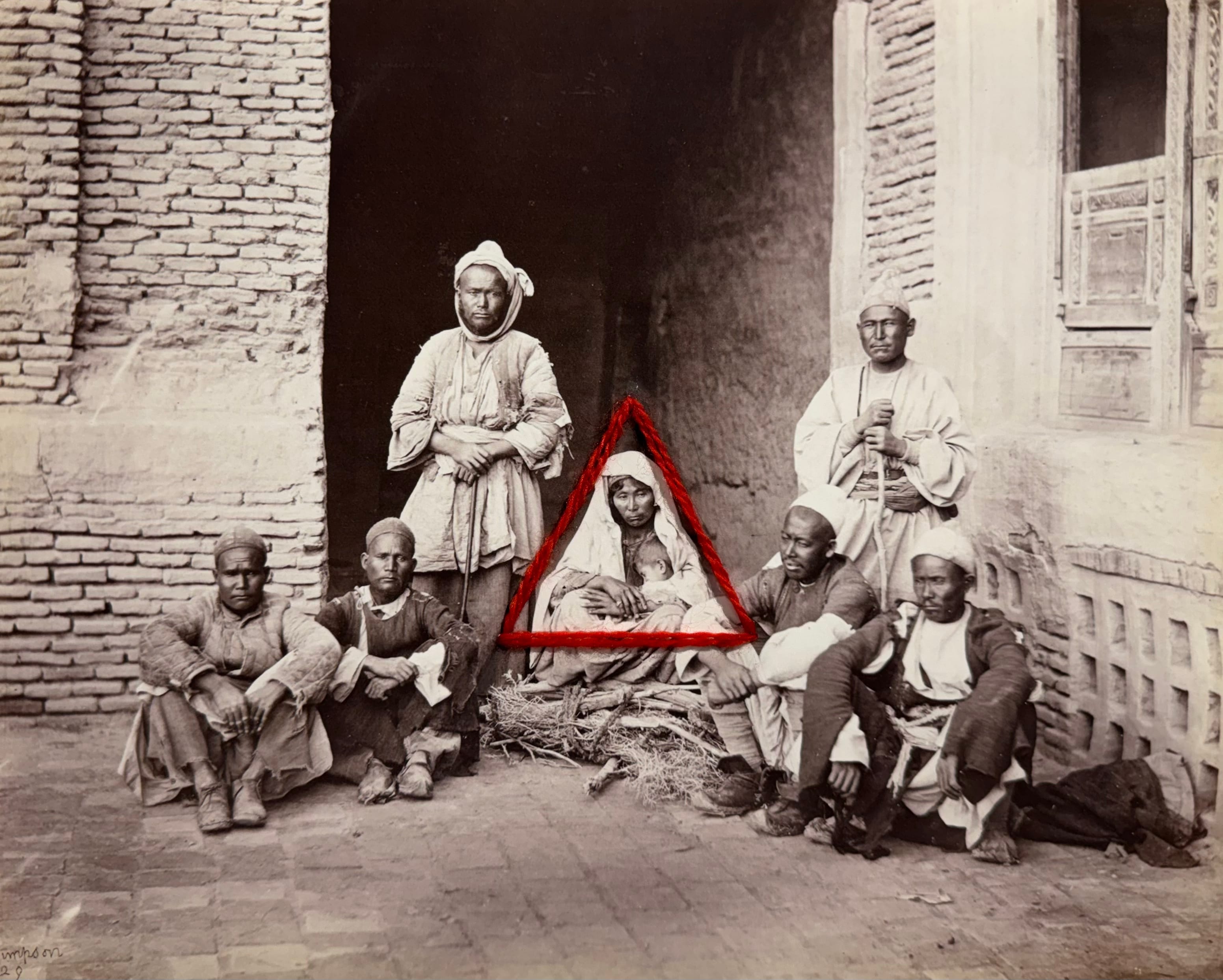 Group of people in traditional clothing seated and standing by a brick wall, with a red triangle drawn around a central seated person.