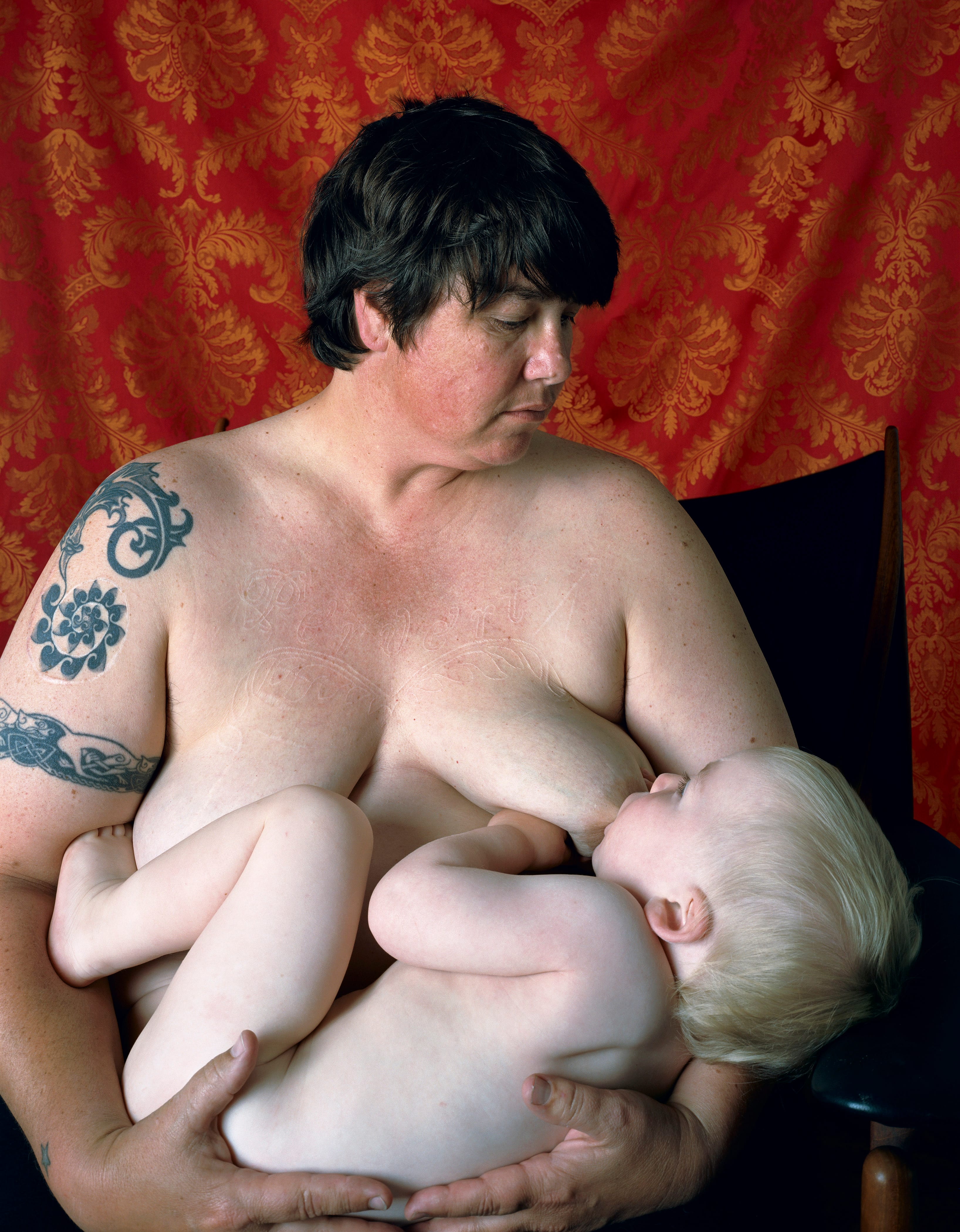 A person with a tattoo breastfeeds a child against a red patterned backdrop.