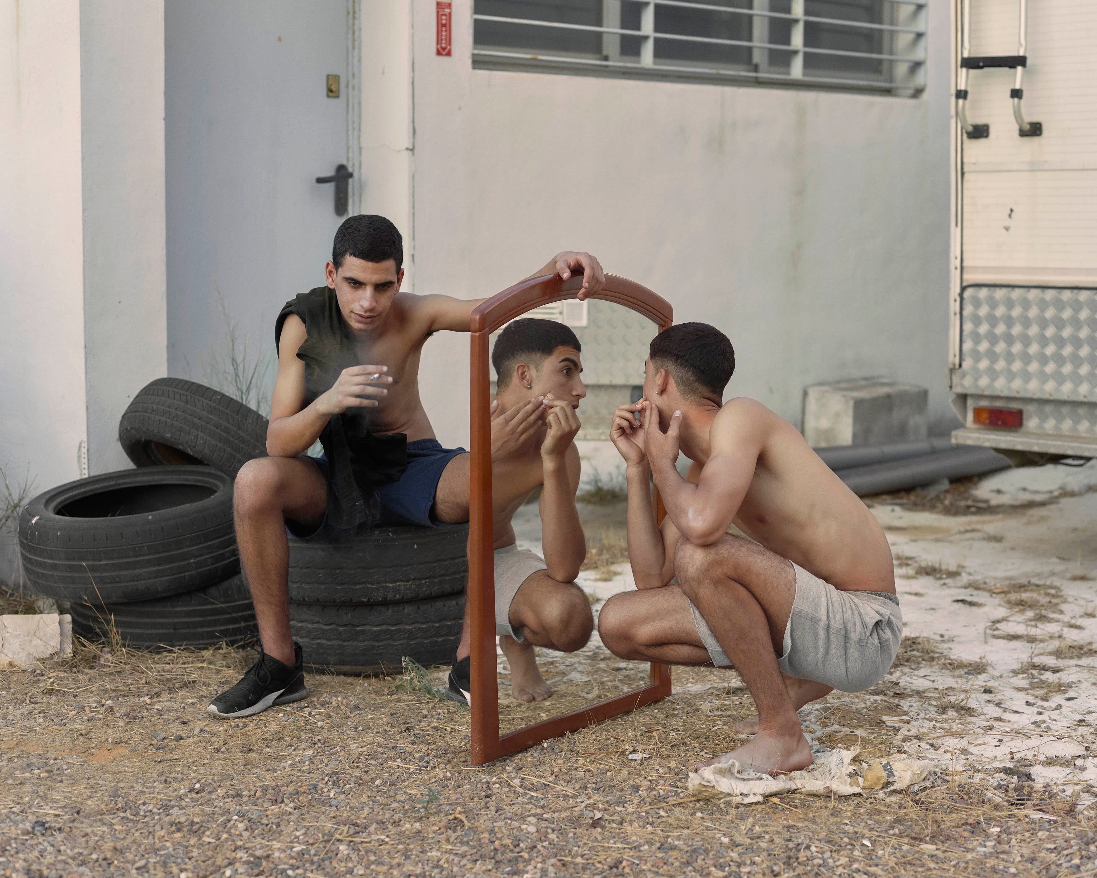 Young boys out on the streets. One boy is grooming in a mirror, while the other boy holds the mirror and looks serious into the camera.