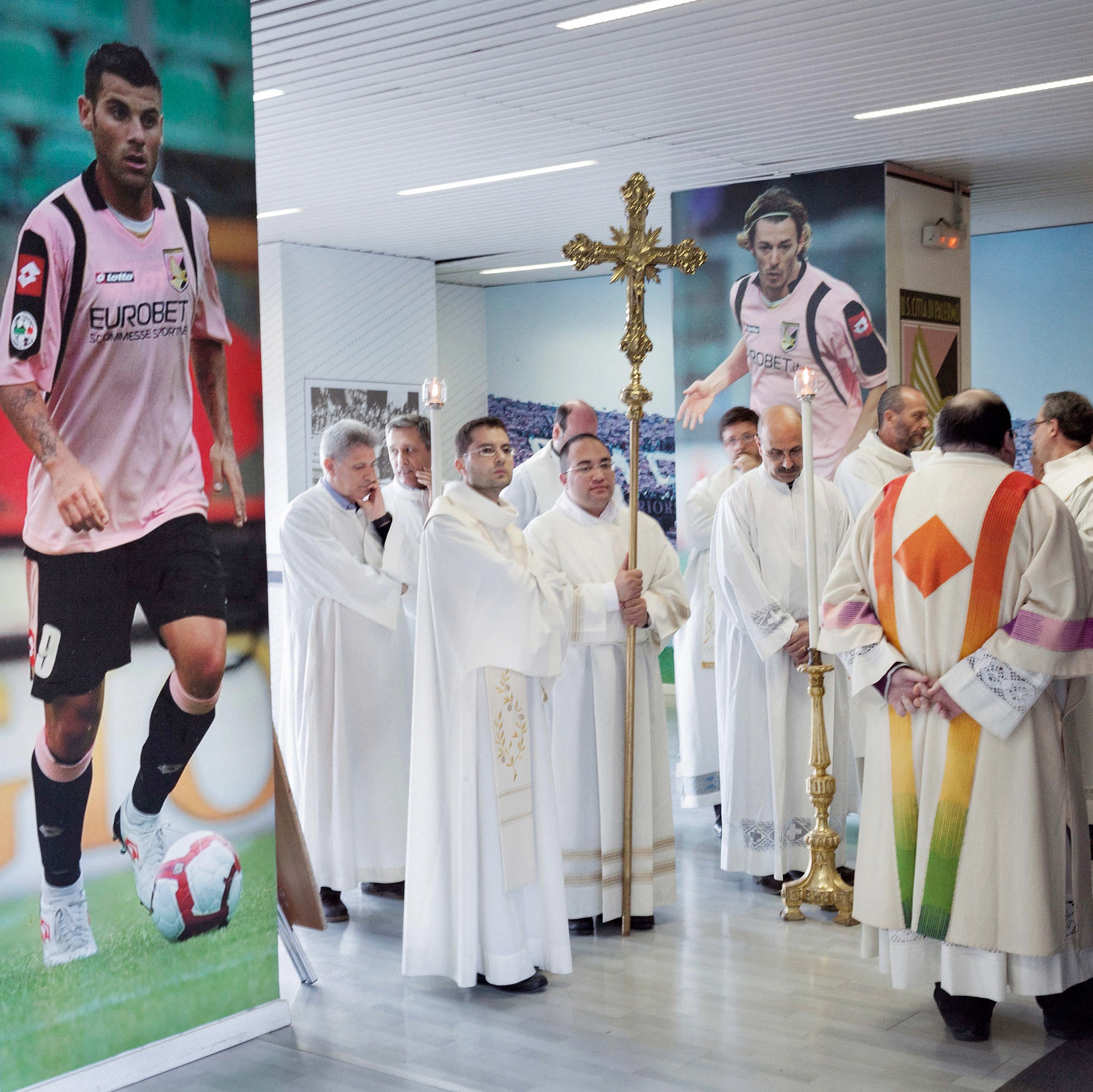 A group of clergy in white robes hold a cross during a procession in a room with large soccer player posters on the walls.