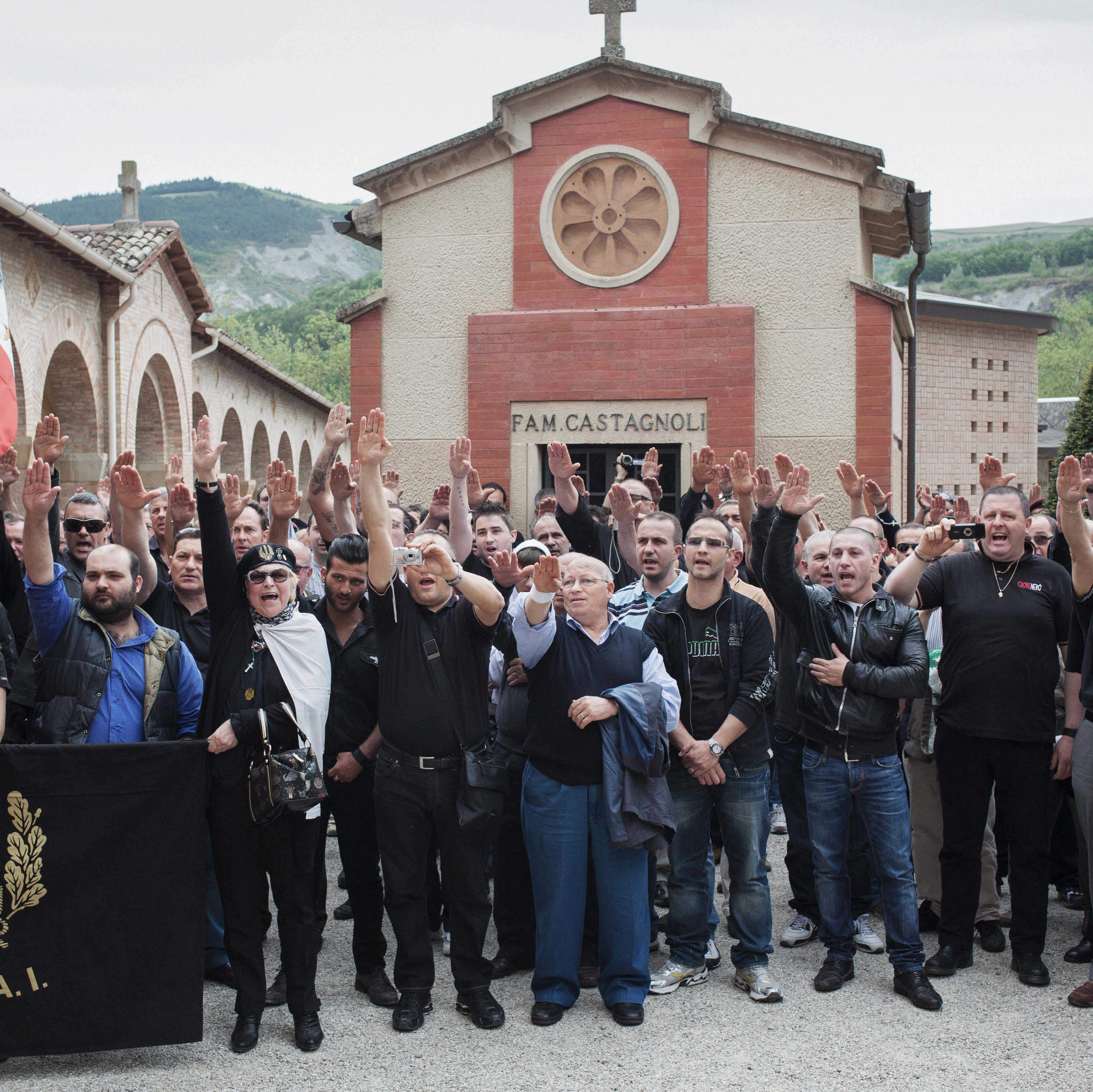 A group of people standing in front of a mausoleum, raising their right arms in a gesture, with a banner visible on the left.