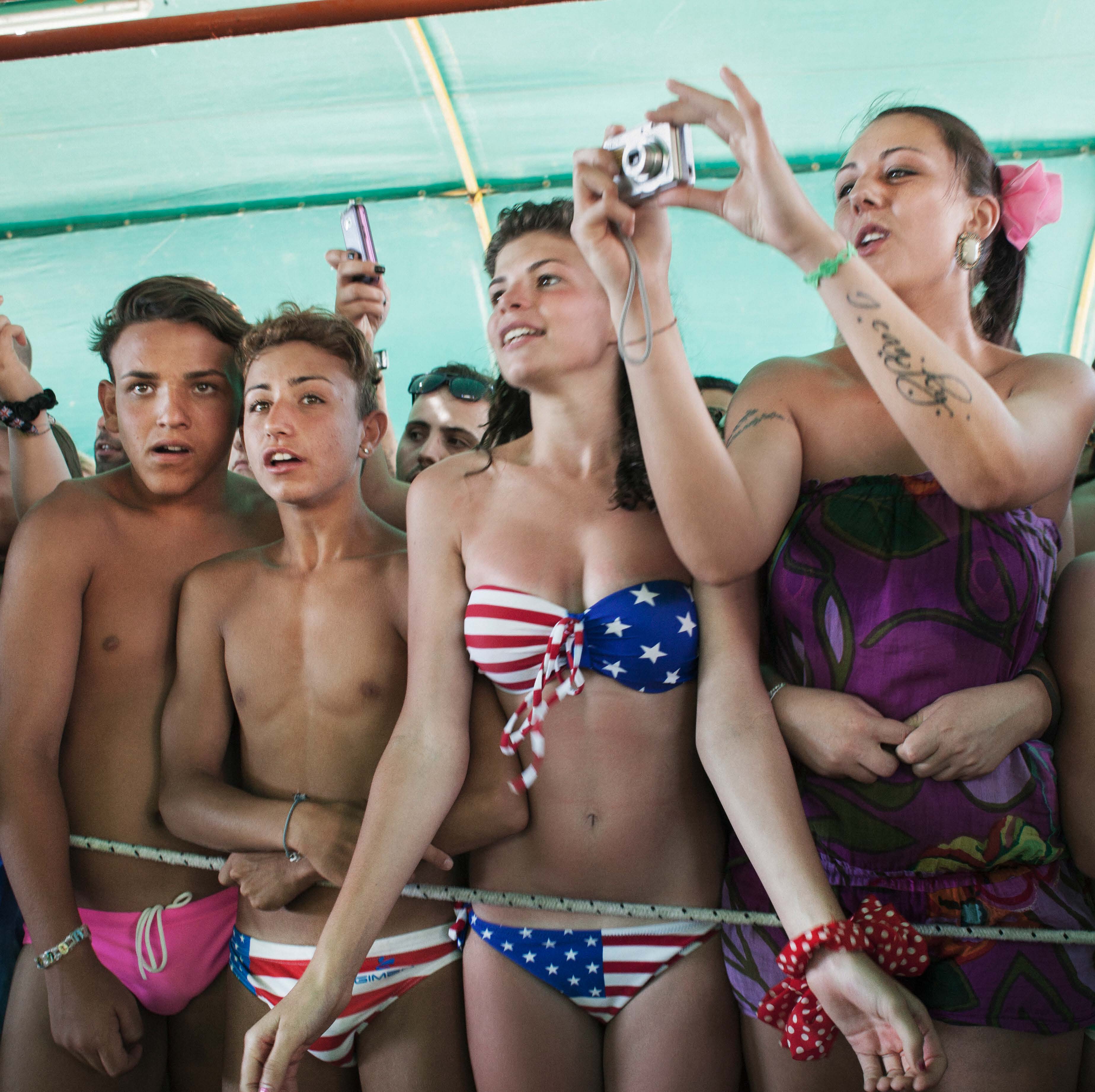 A group of young people in swimsuits, some holding cameras and phones, stand closely together under a tent.