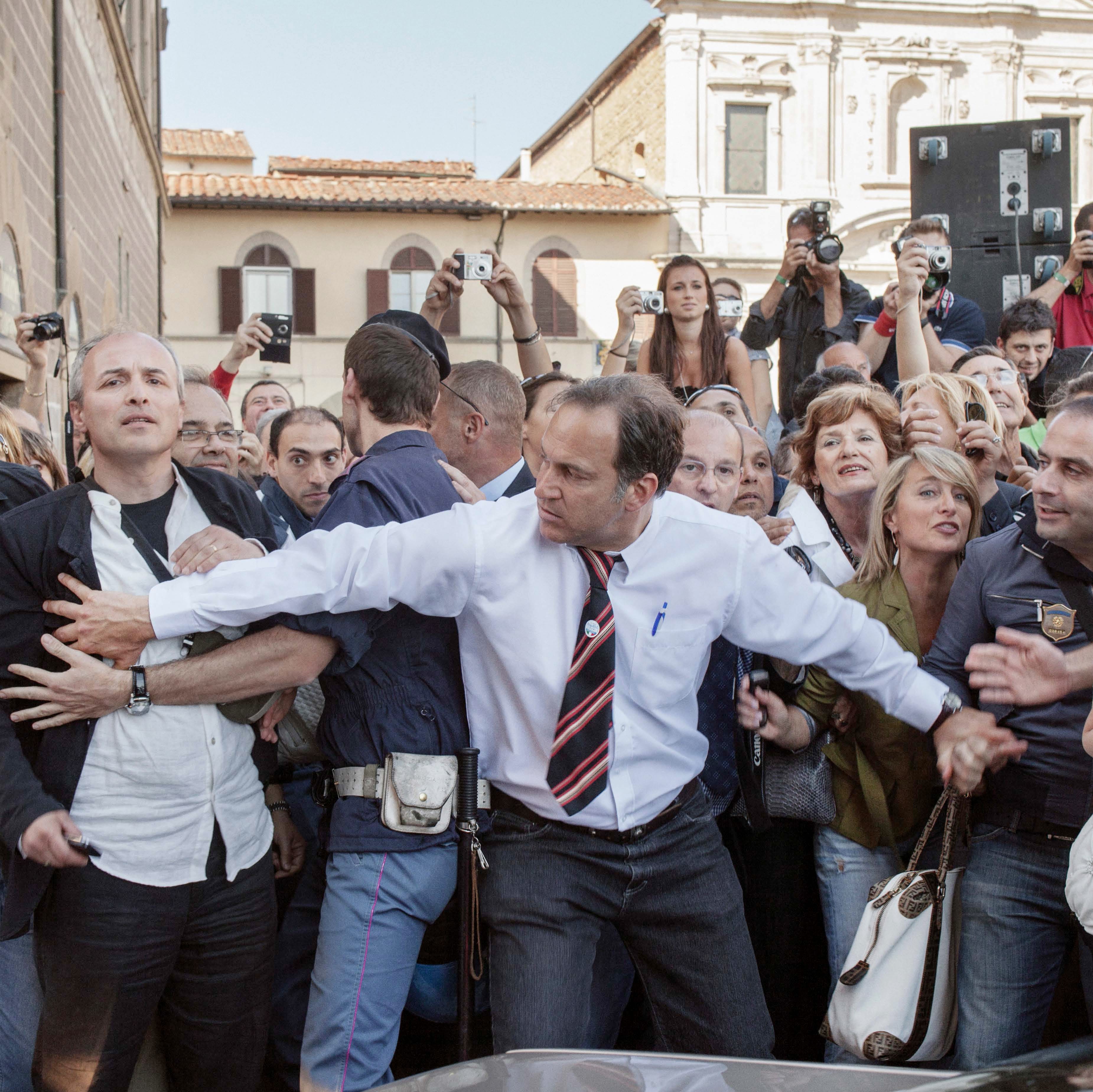 A man in a white shirt and tie holds back a crowd, including photographers, in an outdoor setting with historic buildings in the background.