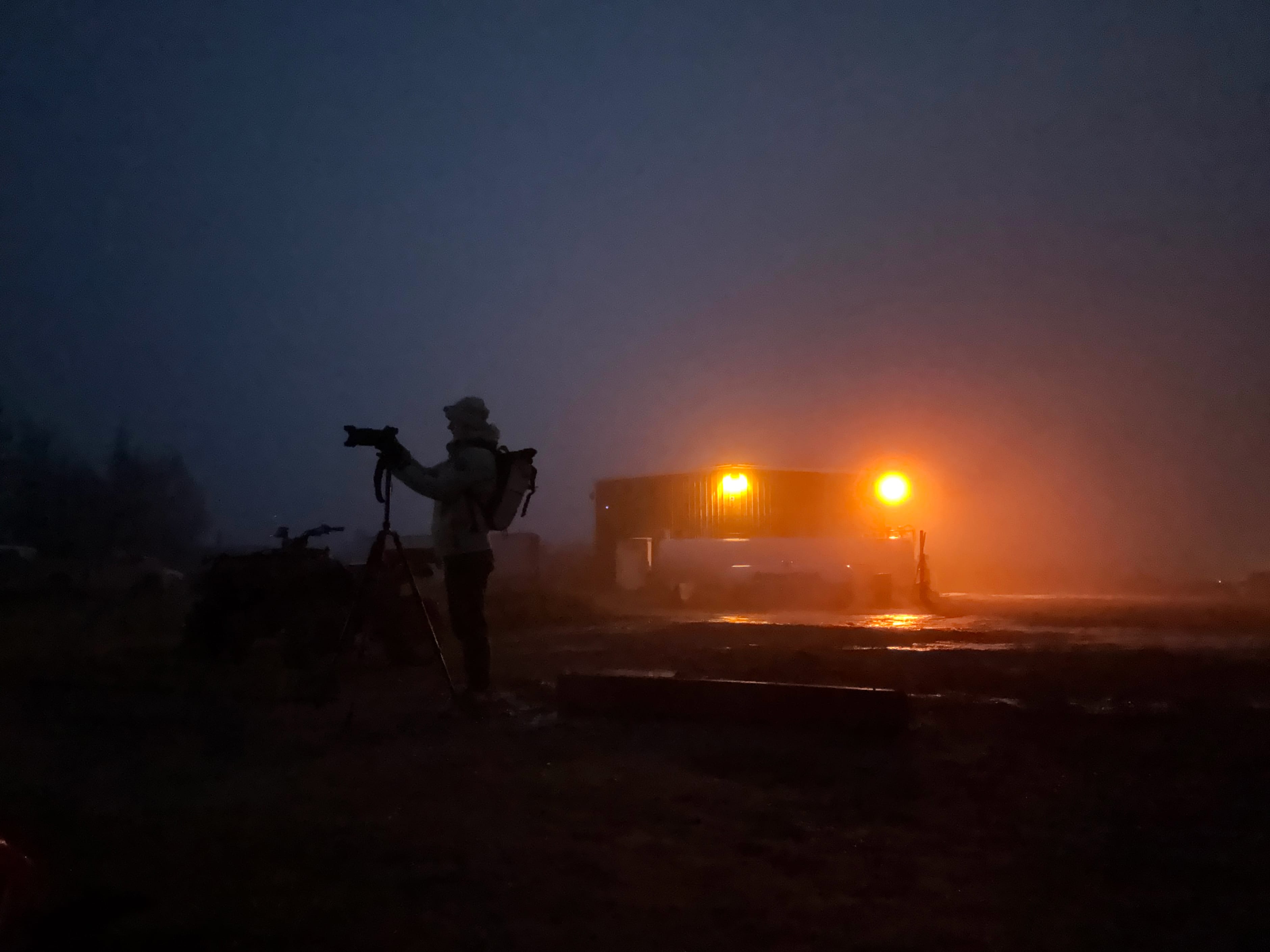 Silhouetted figure with camera at night, facing a building with bright lights in a foggy setting.
