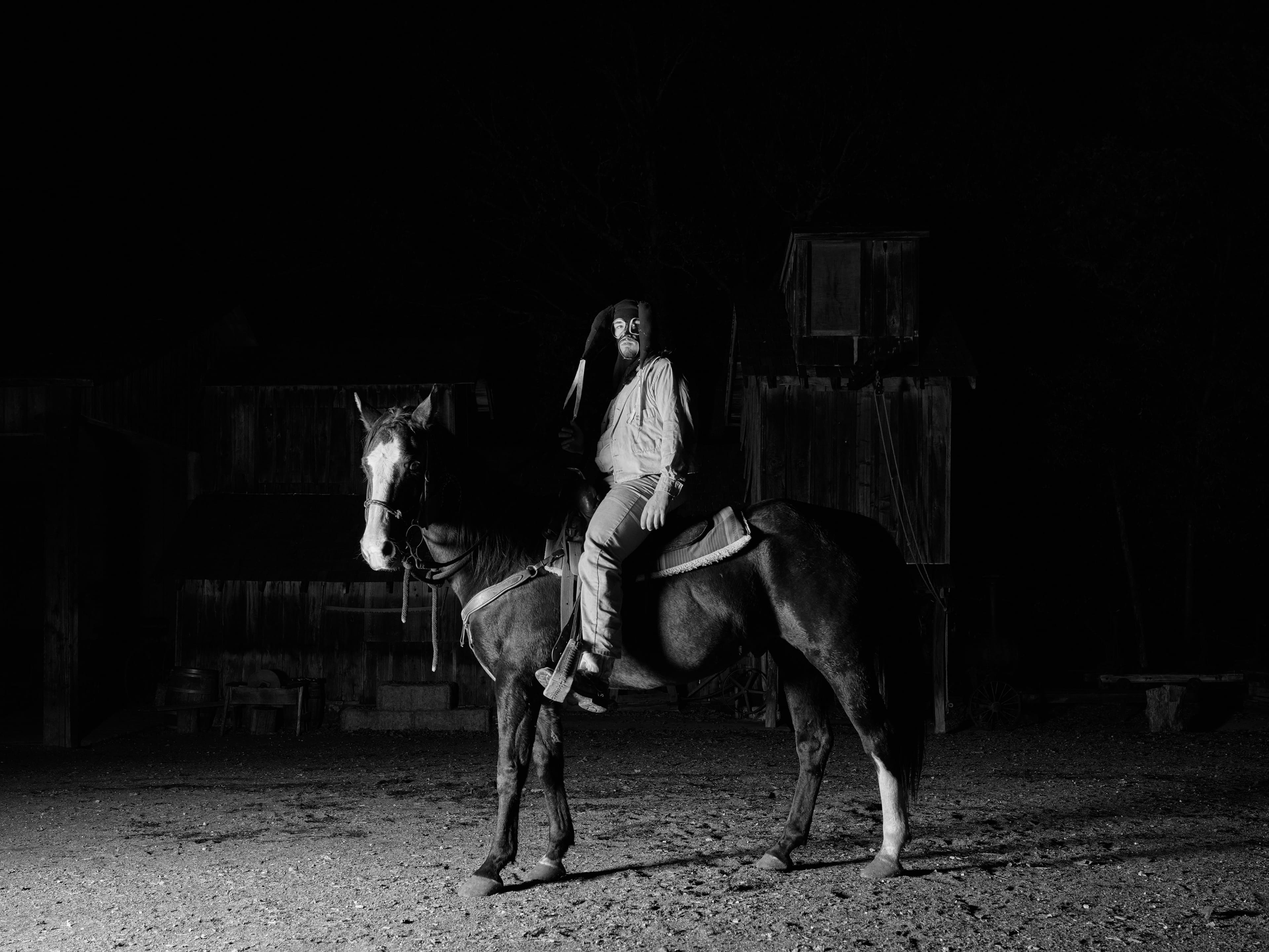 A man in costume, wearing goggles and feathers, sits on a horse in a dimly lit, eerie setting with dark wooden structures in the background.