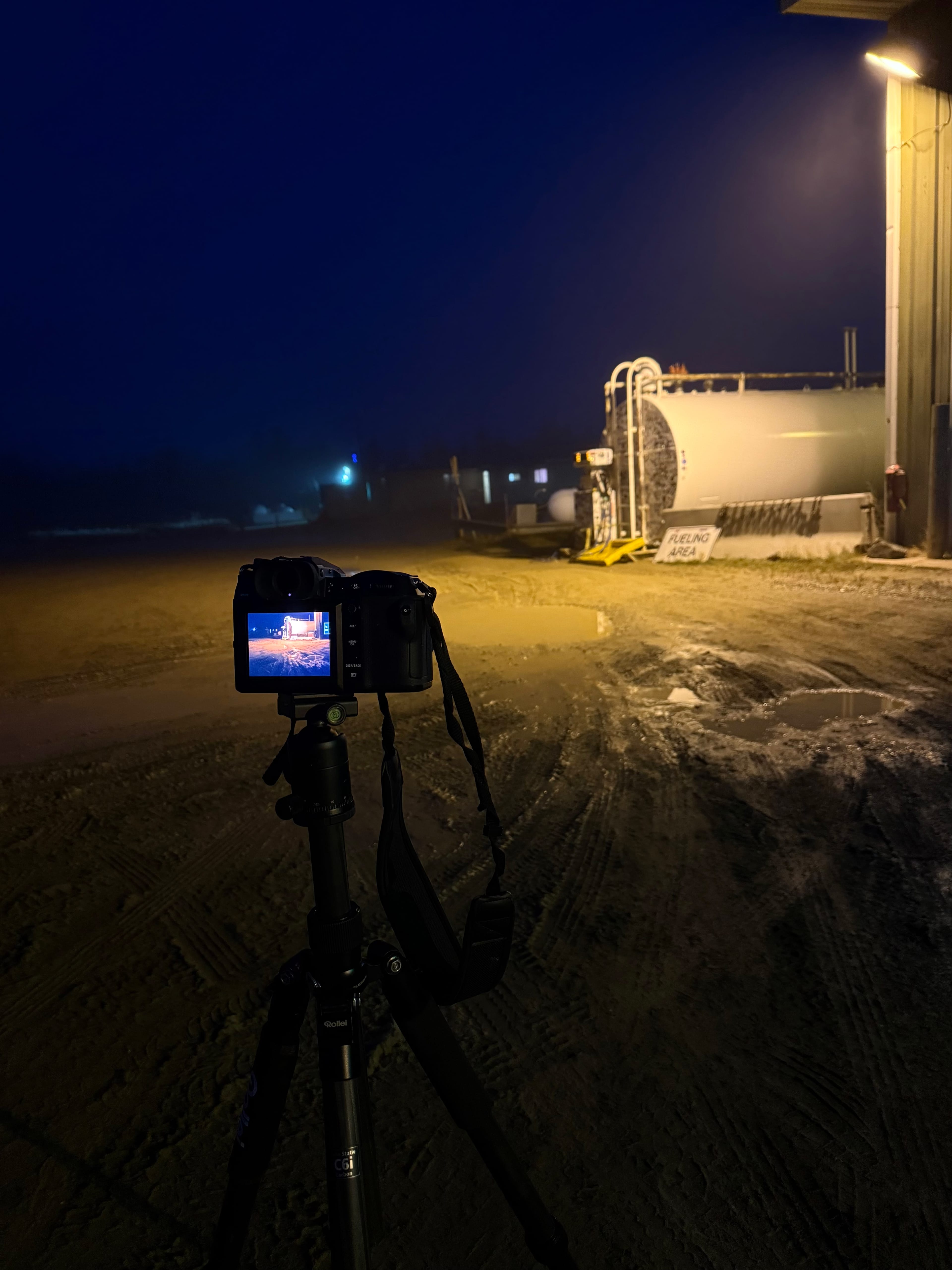 Camera on tripod capturing a dimly lit industrial scene at night, with a large tank and building illuminated by a warm light.