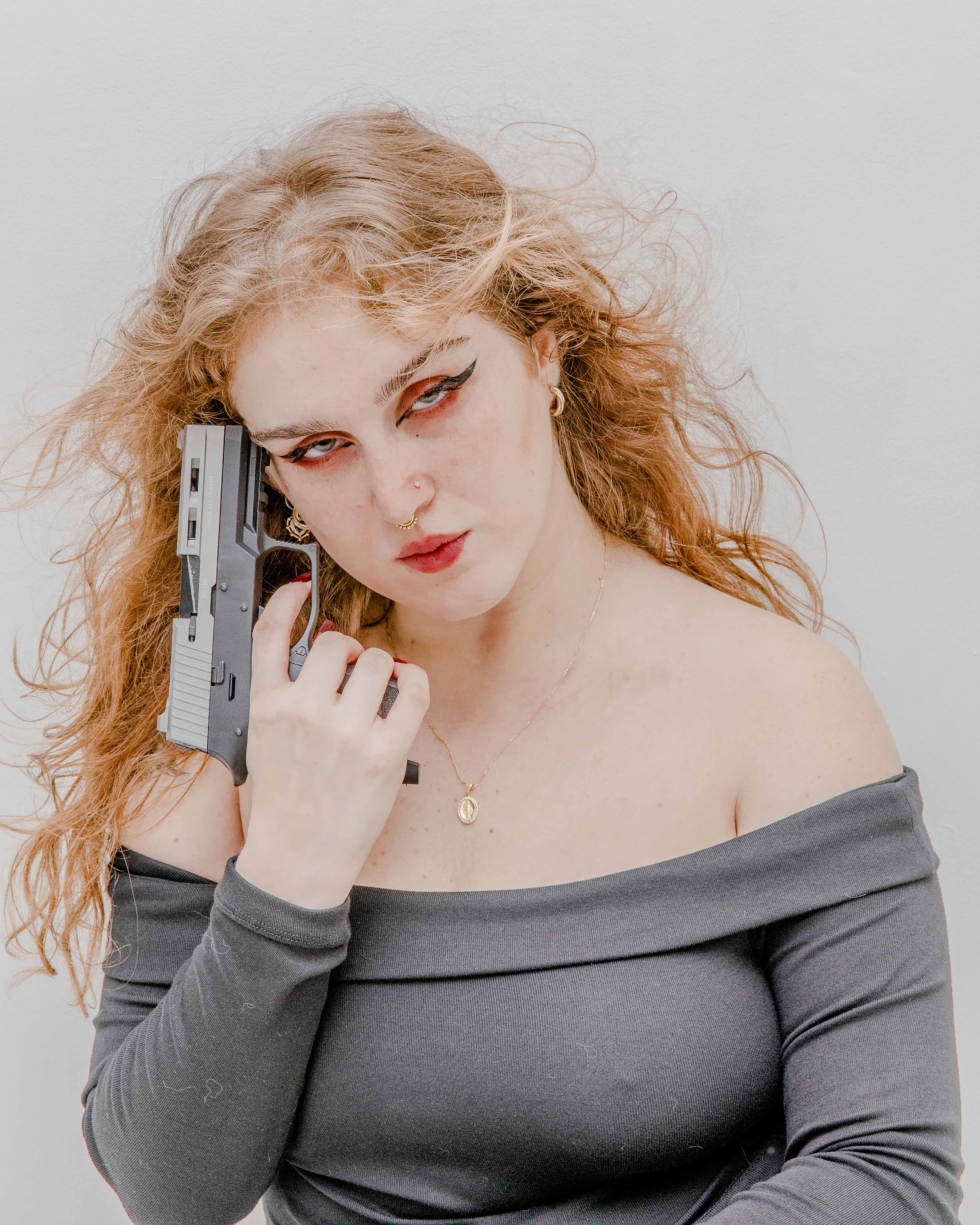 Woman with wavy hair, wearing an off-shoulder top, holds a handgun near her face against a plain background.