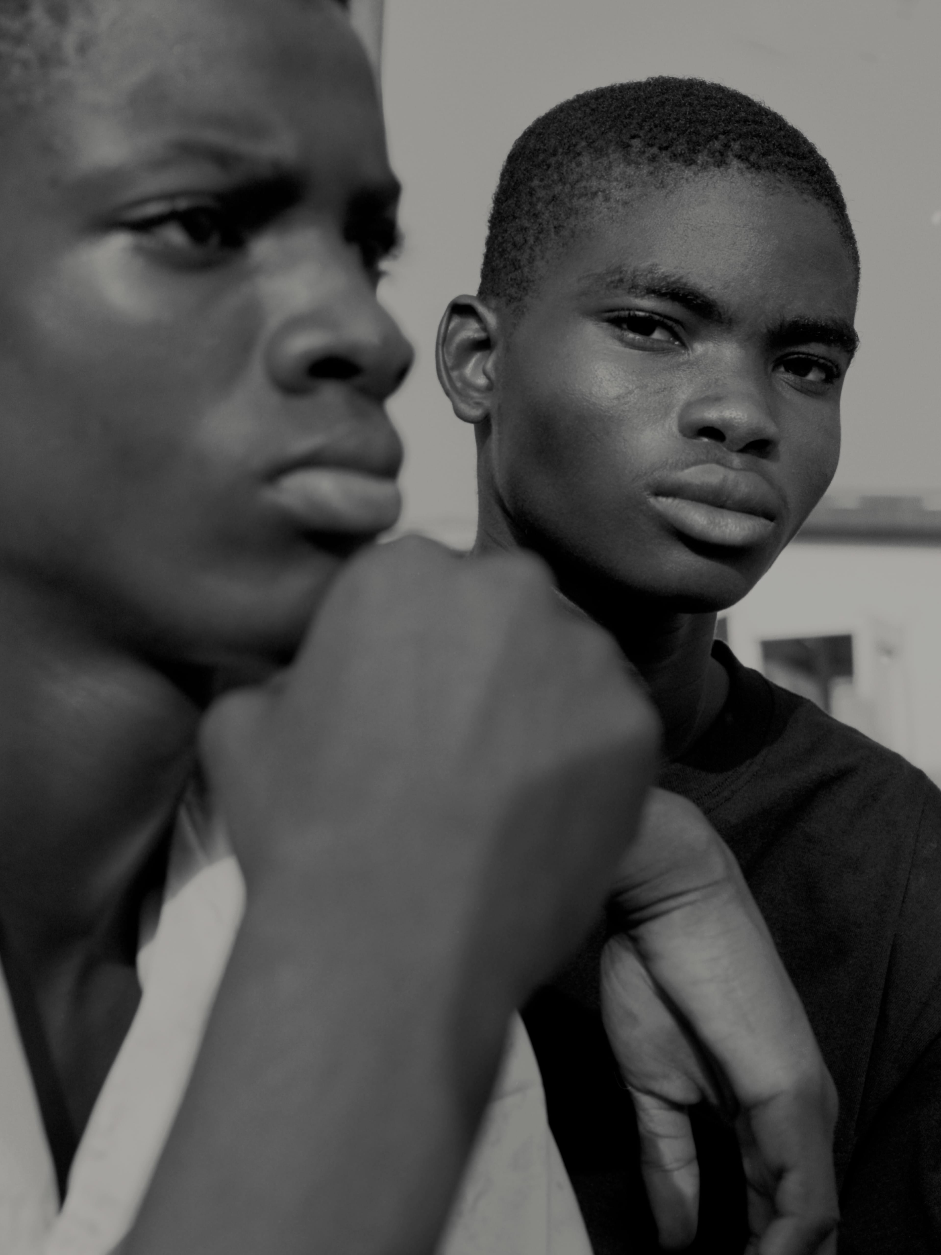 Two young men in a black and white portrait; one in focus with a serious expression, the other blurred in the foreground.