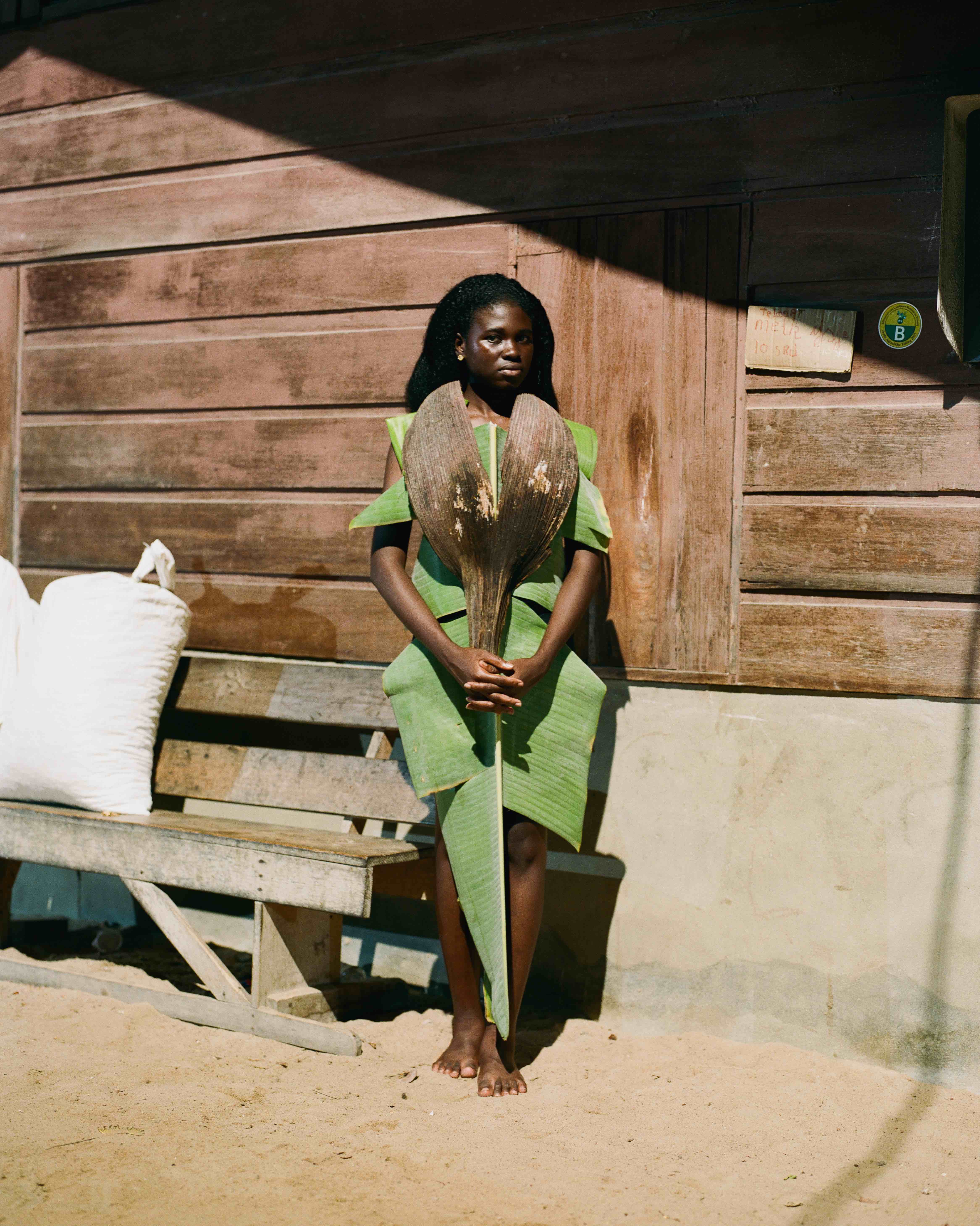 Person in green leaf attire holding a large seed pod, standing against a wooden wall with a shadow overhead.