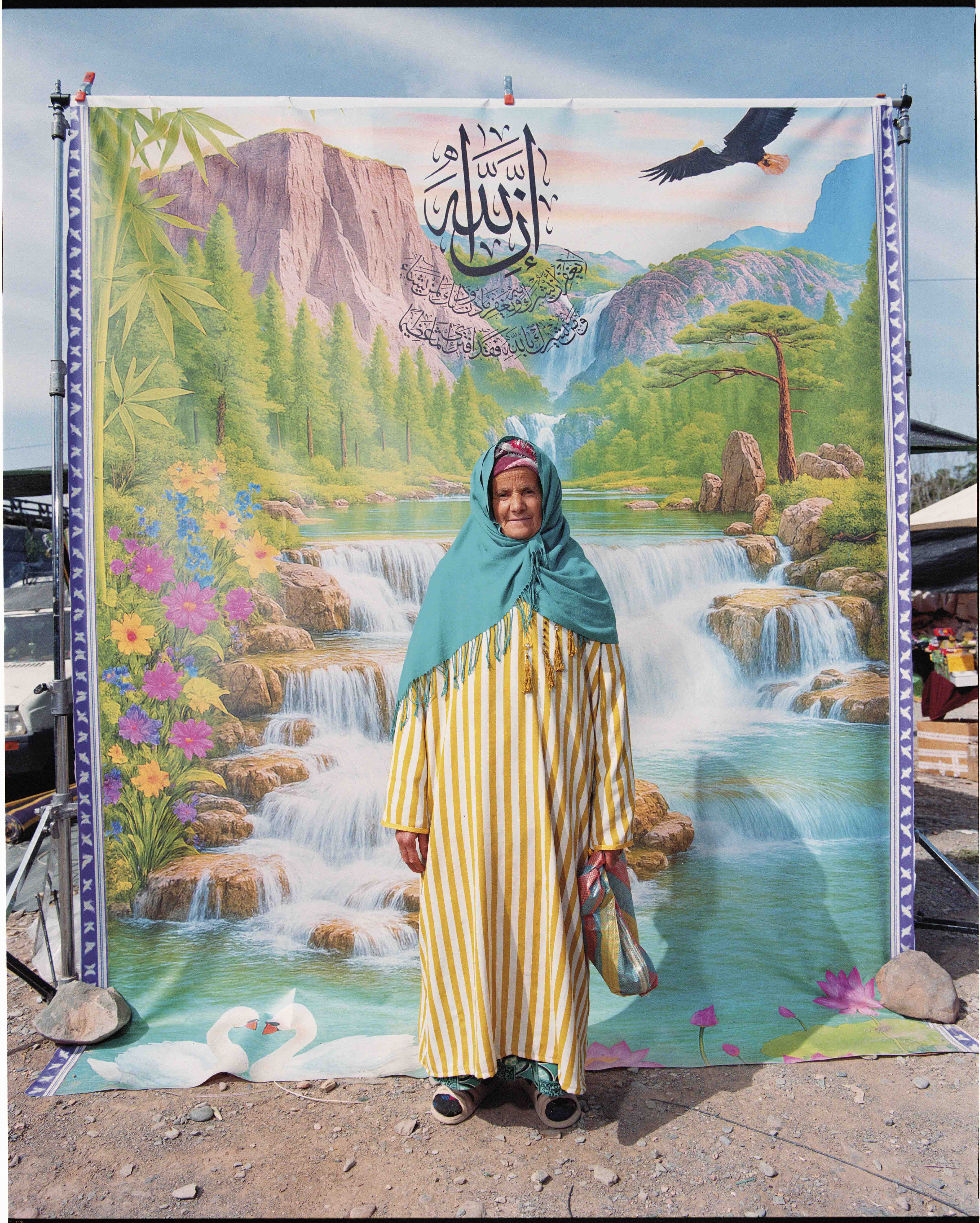 An elderly woman wearing a striped dress and turquoise hijab stands in front of a colorful nature backdrop featuring a waterfall and mountains.