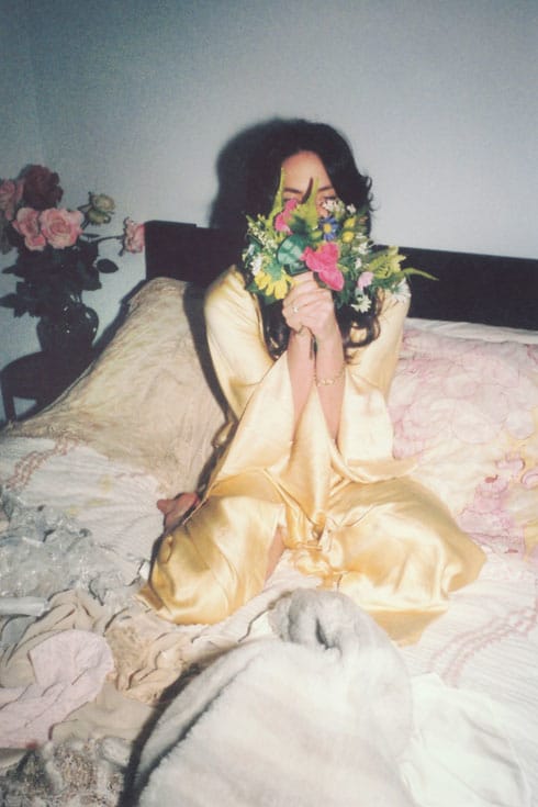 Charlie Hilton sits on a bed in a silk blouse, holding a bouquet of flowers in front of her face.