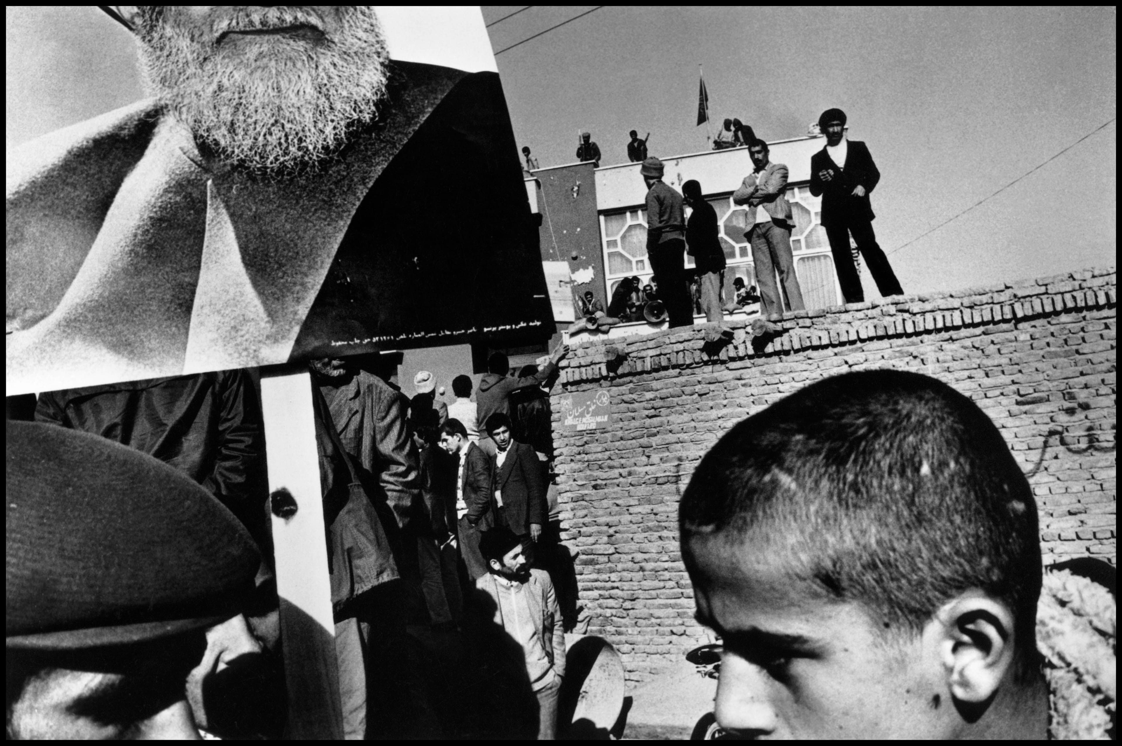 A black-and-white photo showing a crowd near a large portrait of a bearded figure. People stand on a brick wall observing the scene.