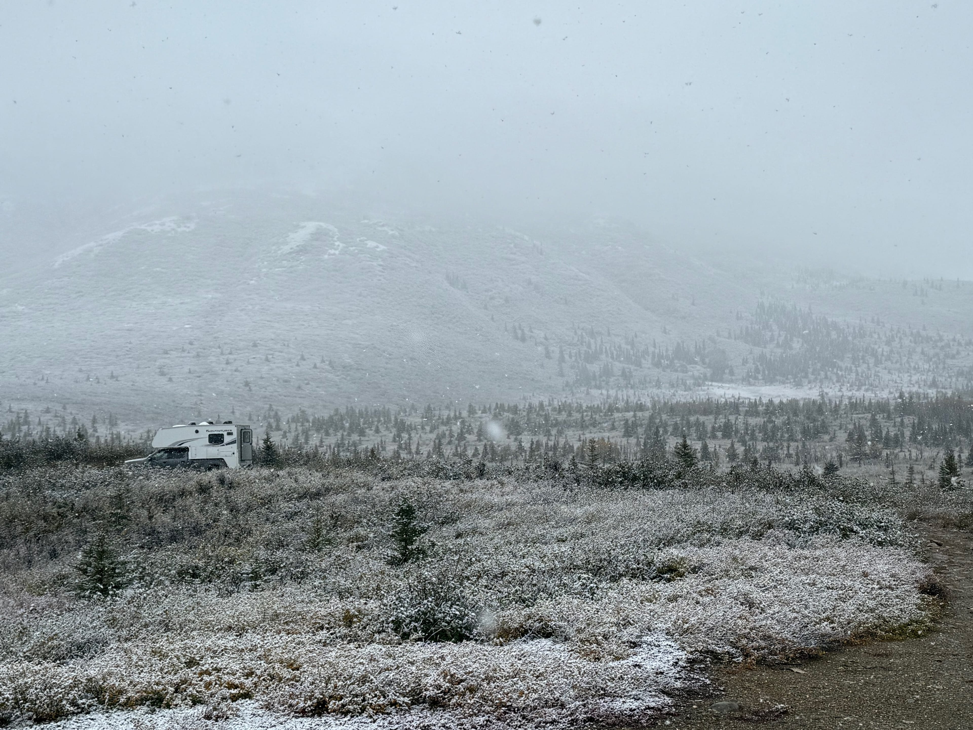 A lone RV parked in a snowy, mountainous landscape under an overcast sky, surrounded by sparse vegetation and distant hills.