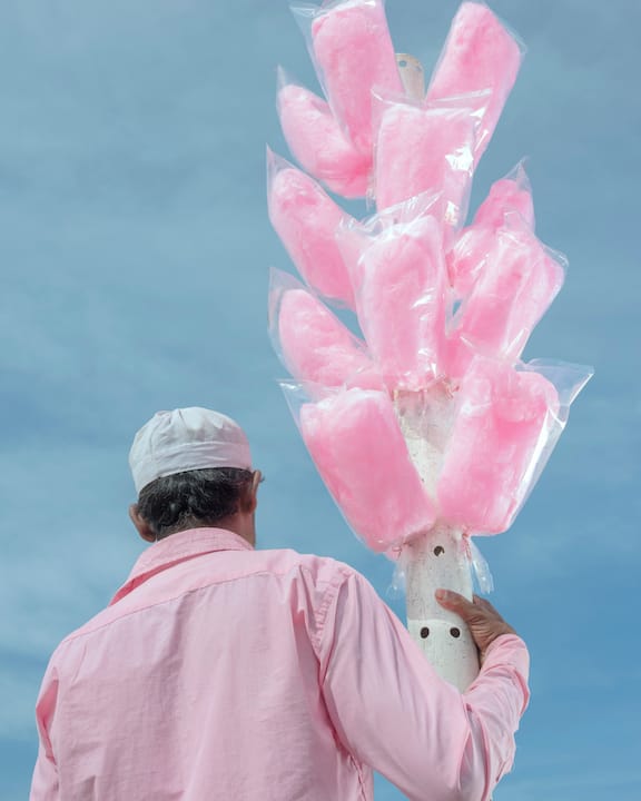A person in a pink shirt and cap holds a pole with multiple bags of pink cotton candy against a blue sky.