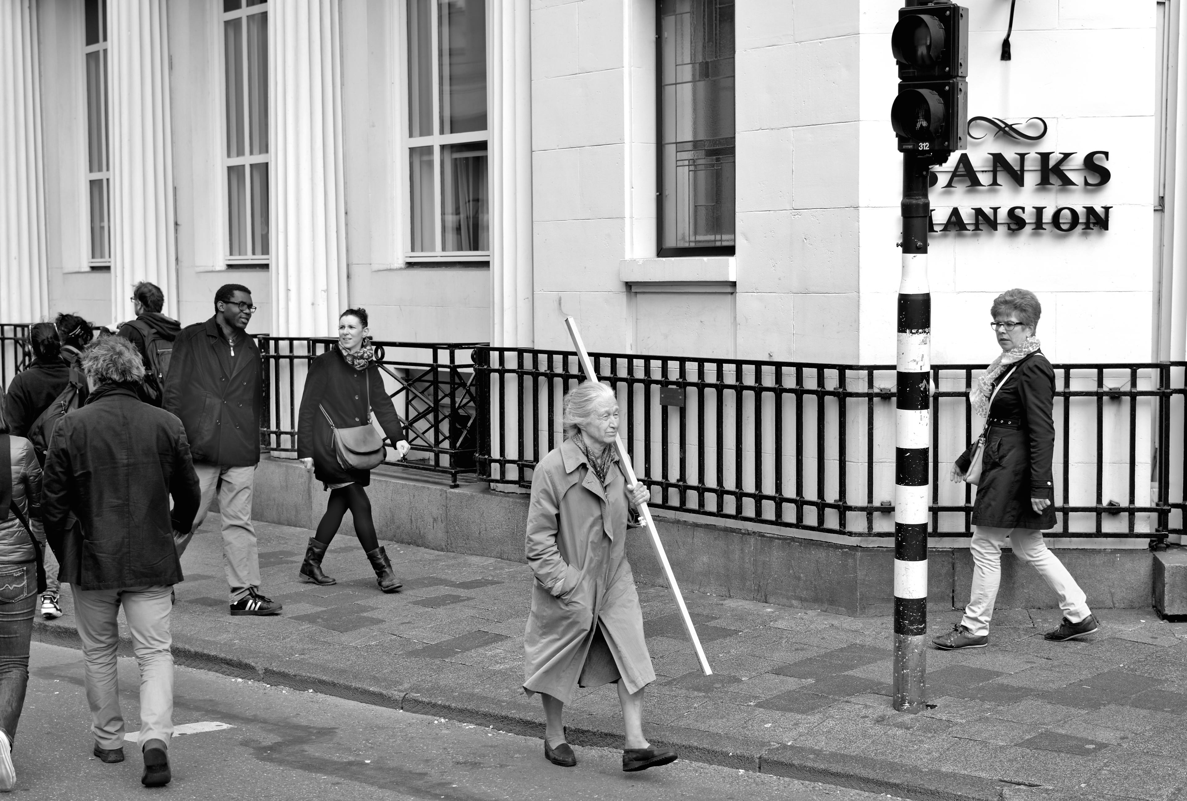 Black and white image of a older woman walking the street with a pole in her hand