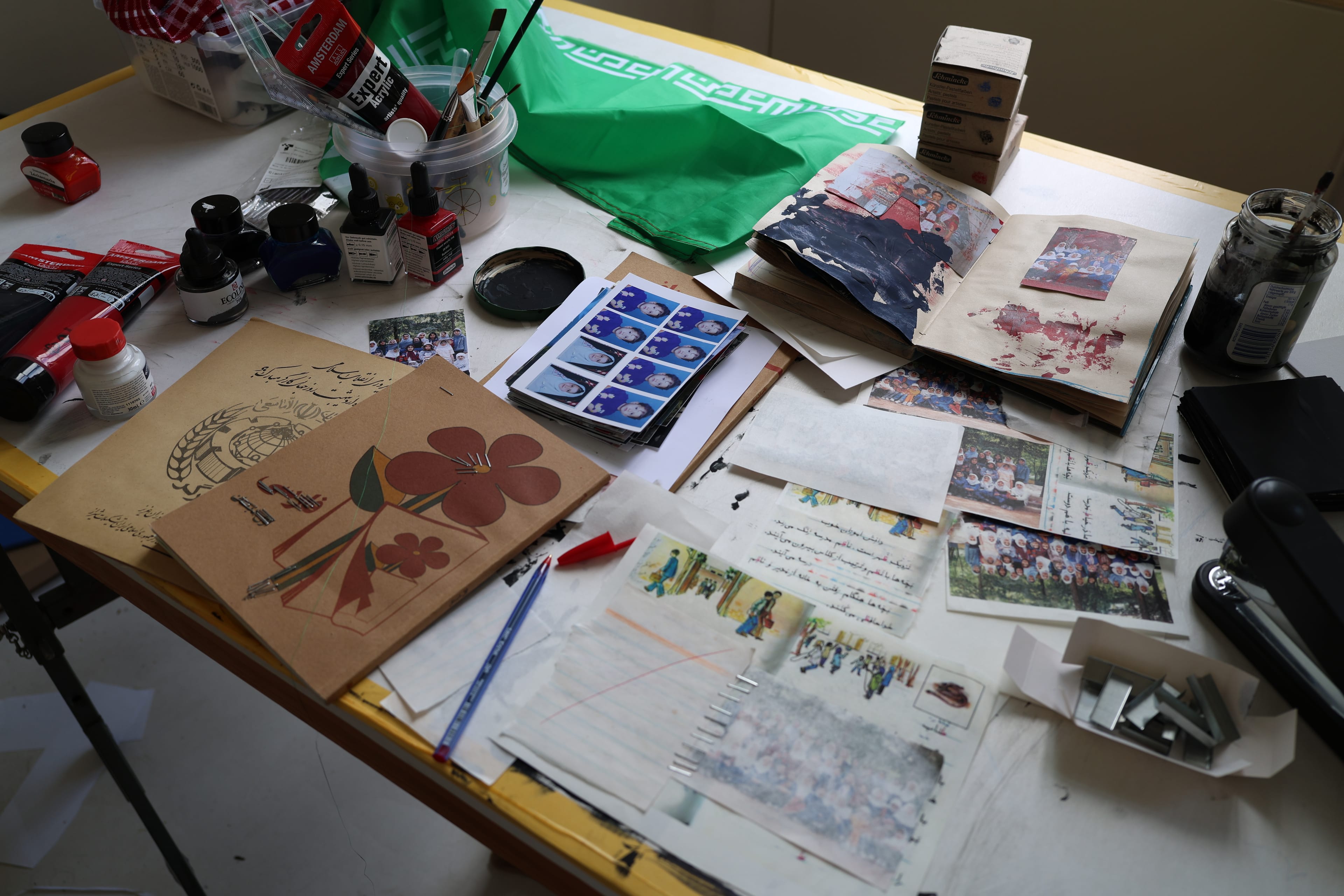 Art supplies and various papers with photos and text are scattered on a cluttered table, with books, jars, and stationery visible.