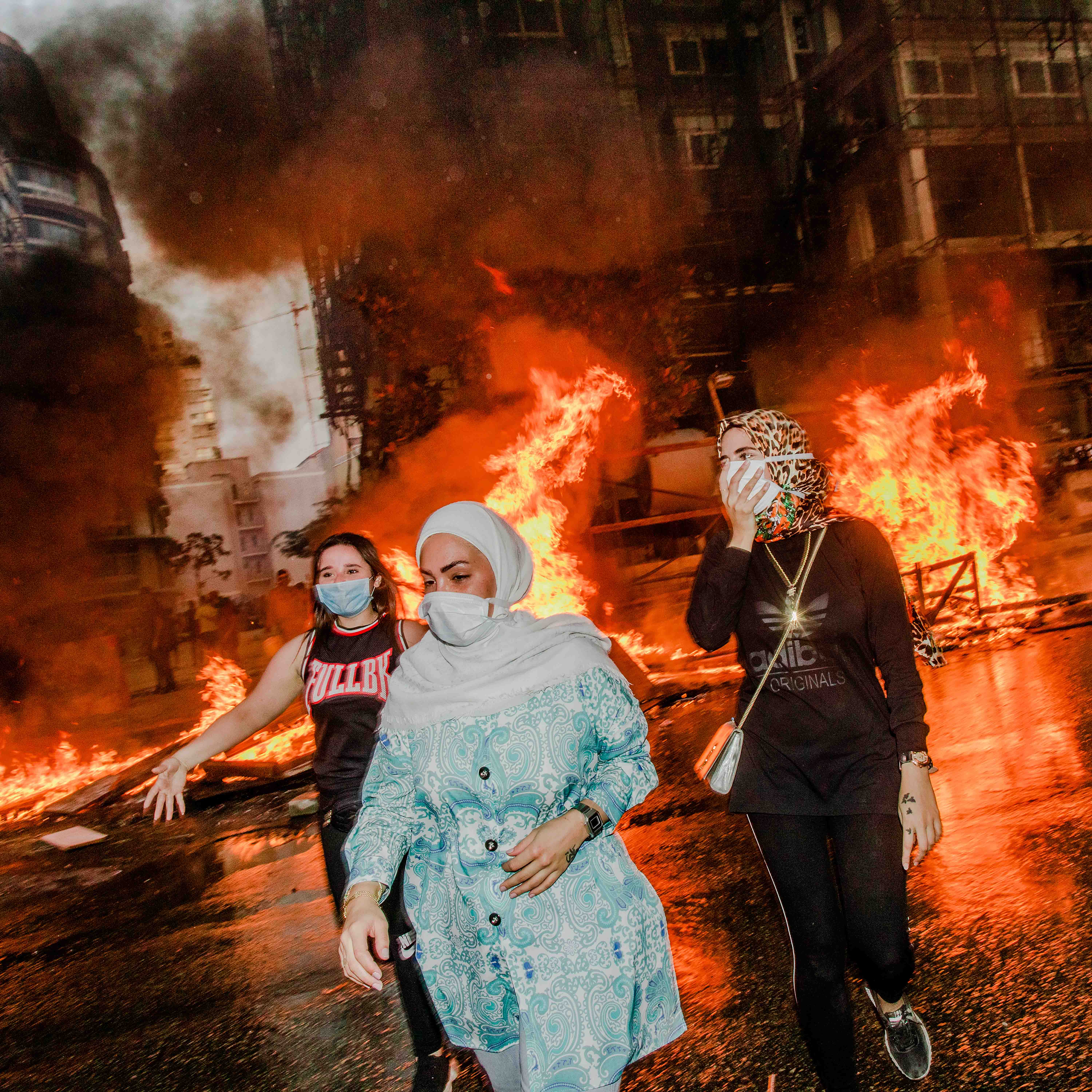Three women in masks walk past a large fire on a city street at night, with flames and smoke rising behind them.