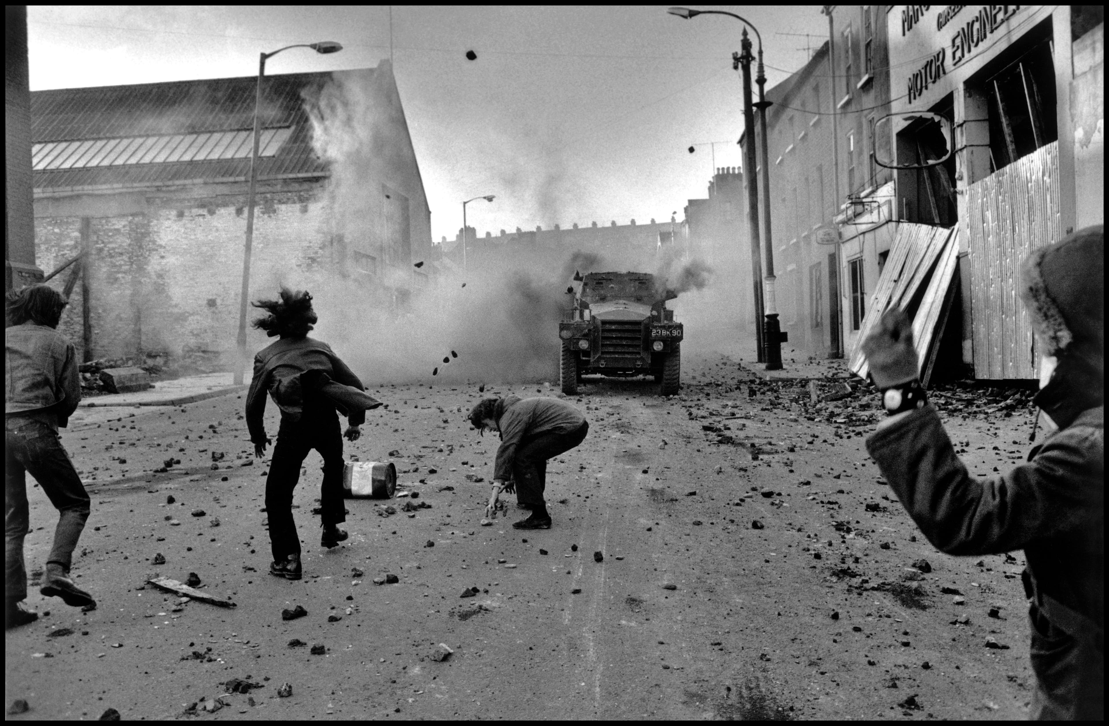 Protest scene on a debris-littered street with people hurling objects at an armored vehicle amid smoke and damaged buildings.