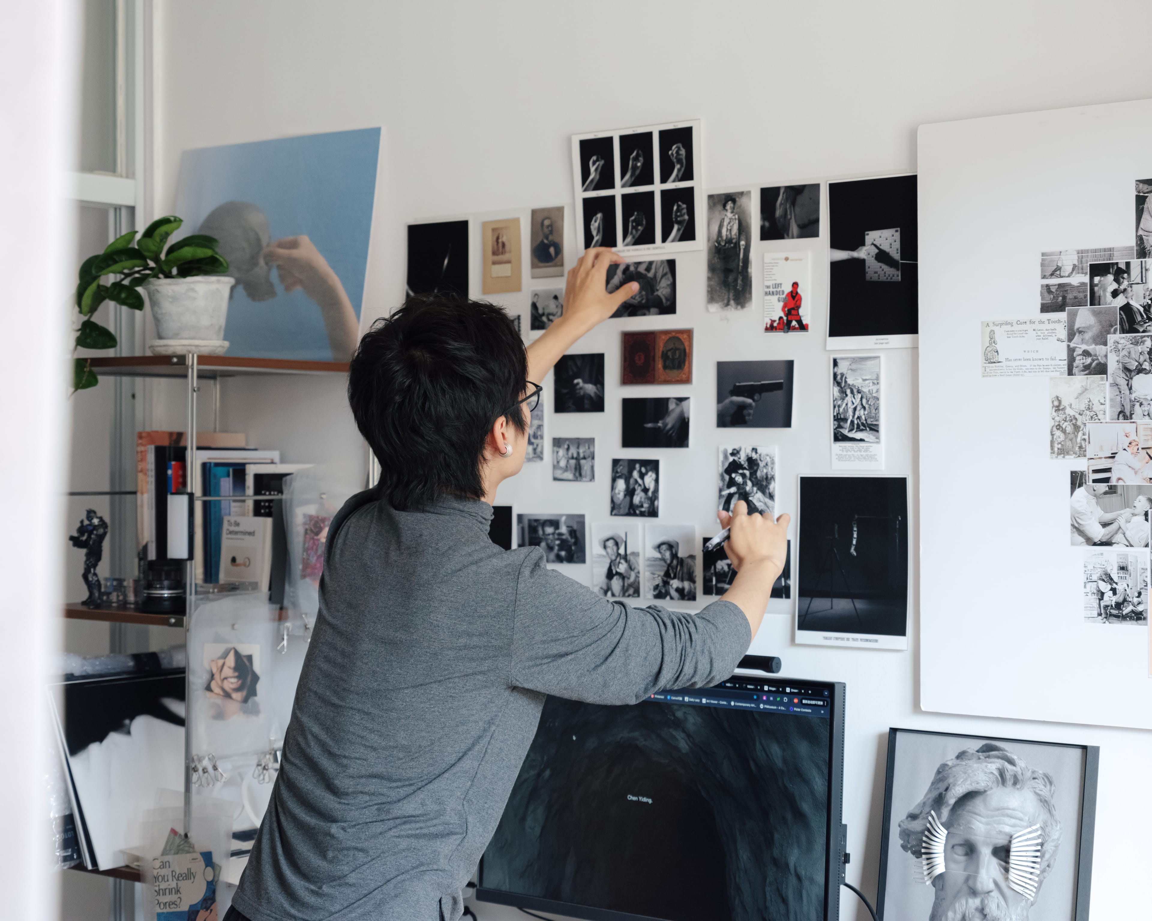 A person arranges photos and artwork on a wall in an art-filled studio, featuring a monitor and various visual elements.