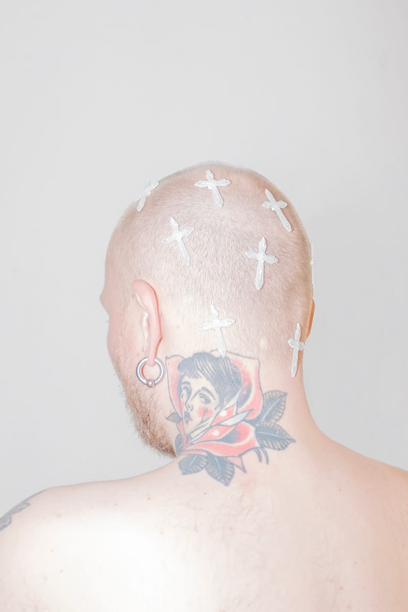 Bald person with hoop earring, cross bandages on head, and a colorful tattoo of a face surrounded by leaves on the neck.