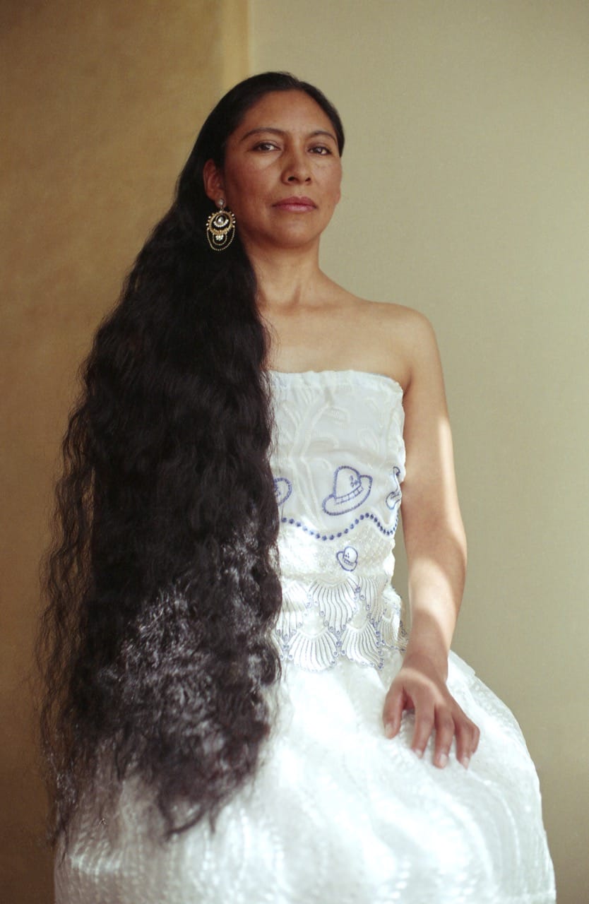 A woman with long black hair and a strapless white dress sits against a light background, wearing a decorative earring.