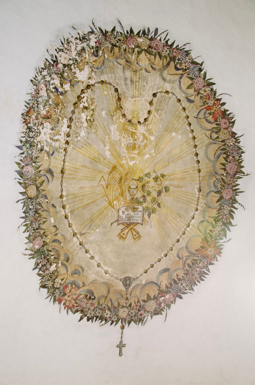 An ornate, oval religious painting with floral border, central sacred symbols, and a rosary with a cross hanging at the bottom.