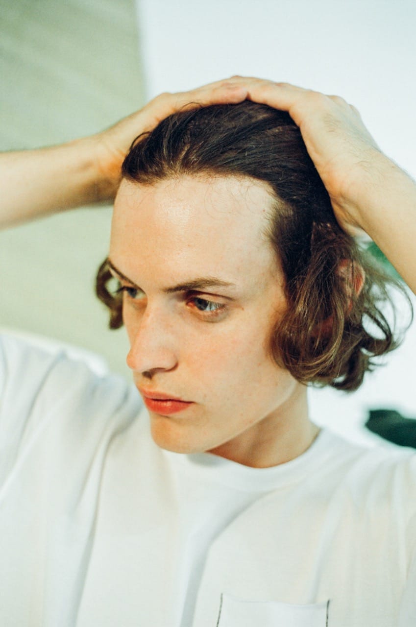 Person with medium-length hair in a white shirt, holding their head with both hands, looking to the side.